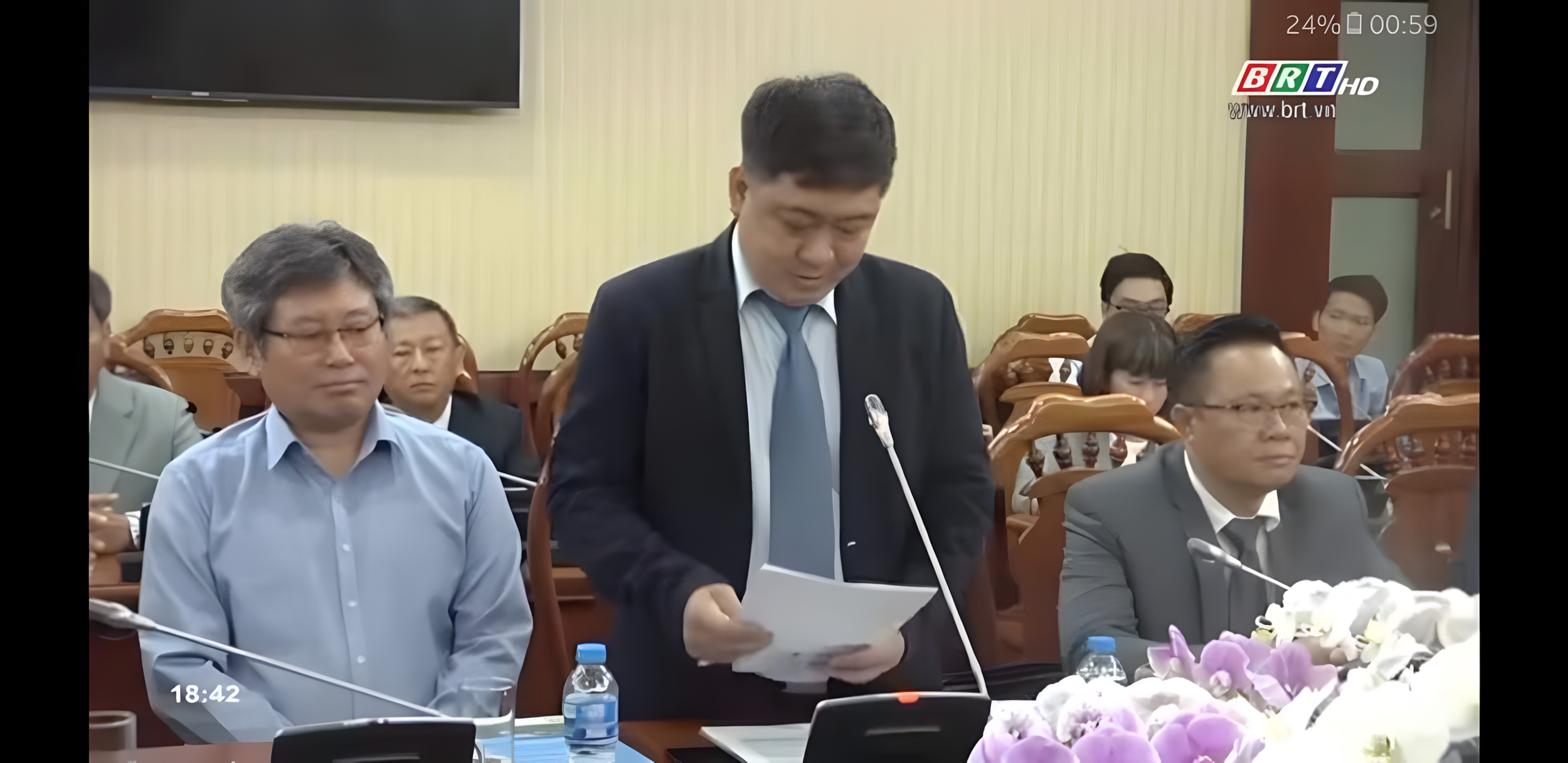 A formal meeting with several Asian attendees, many in suits, seated at a table with microphones, documents, water bottles, and flower arrangements, in a conference room with a television screen and beige curtains.