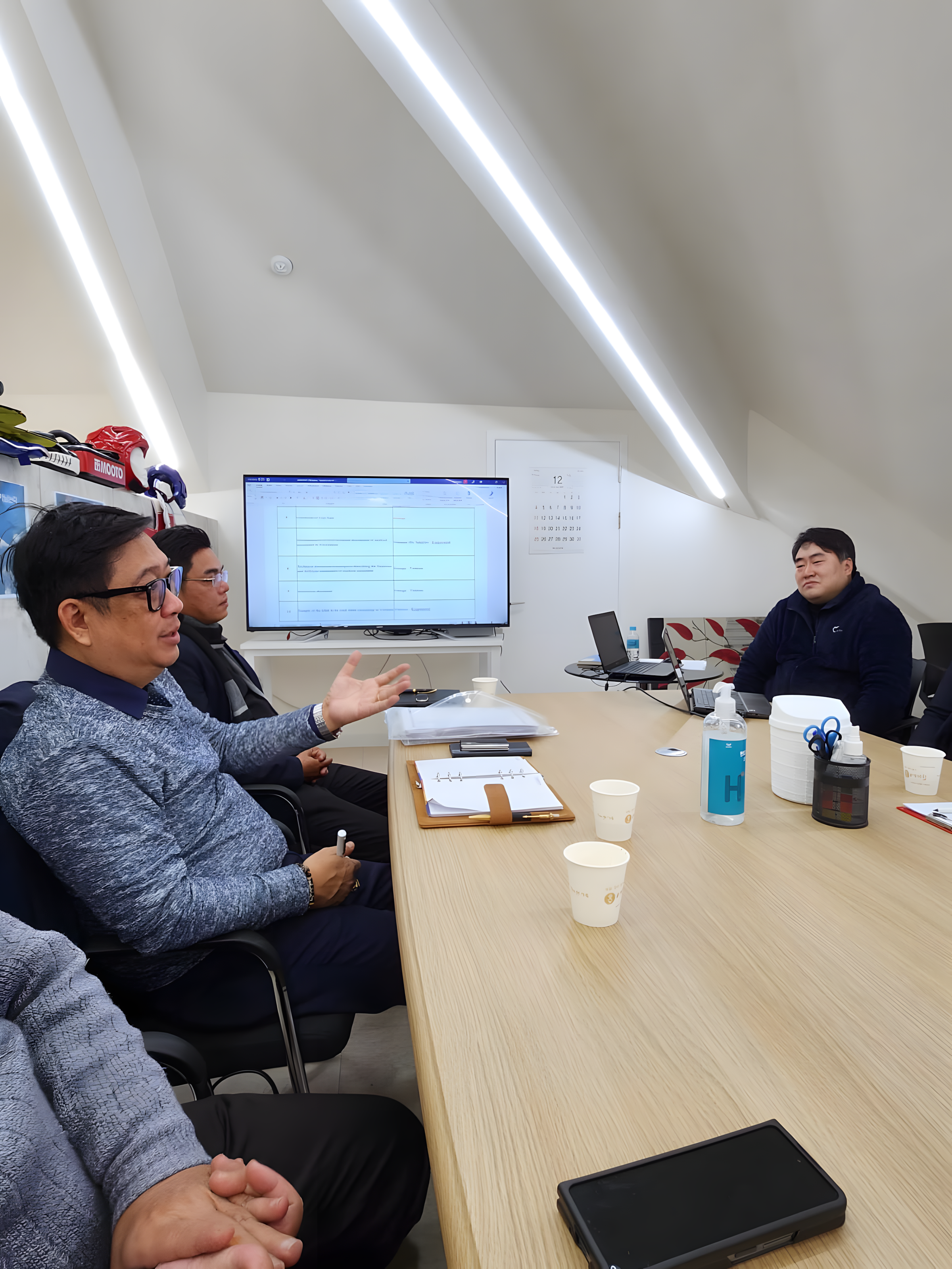 Business meeting in a conference room with four people, a large monitor displaying a spreadsheet, notebooks, disposable cups, hand sanitizer, and laptops on the table.
