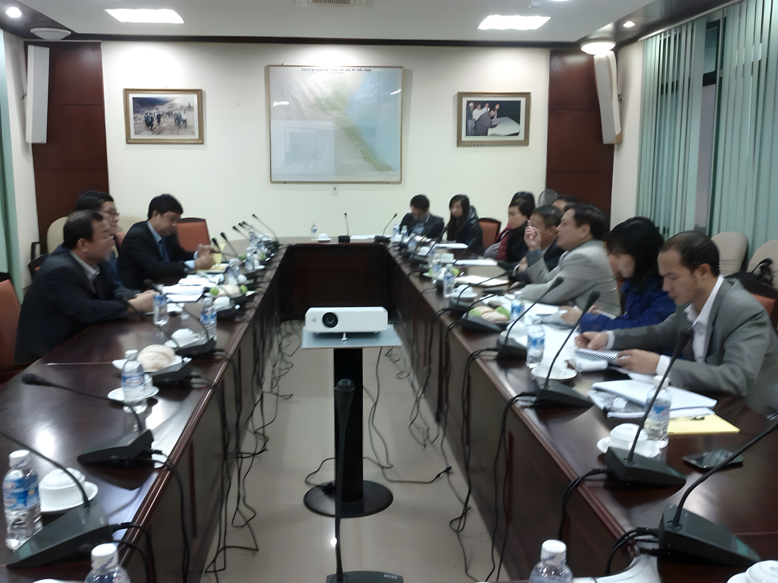 A group of professionals in business attire attending a meeting in a conference room, seated around a large U-shaped table with microphones, water bottles, and documents, with a projector on a stand in the center.