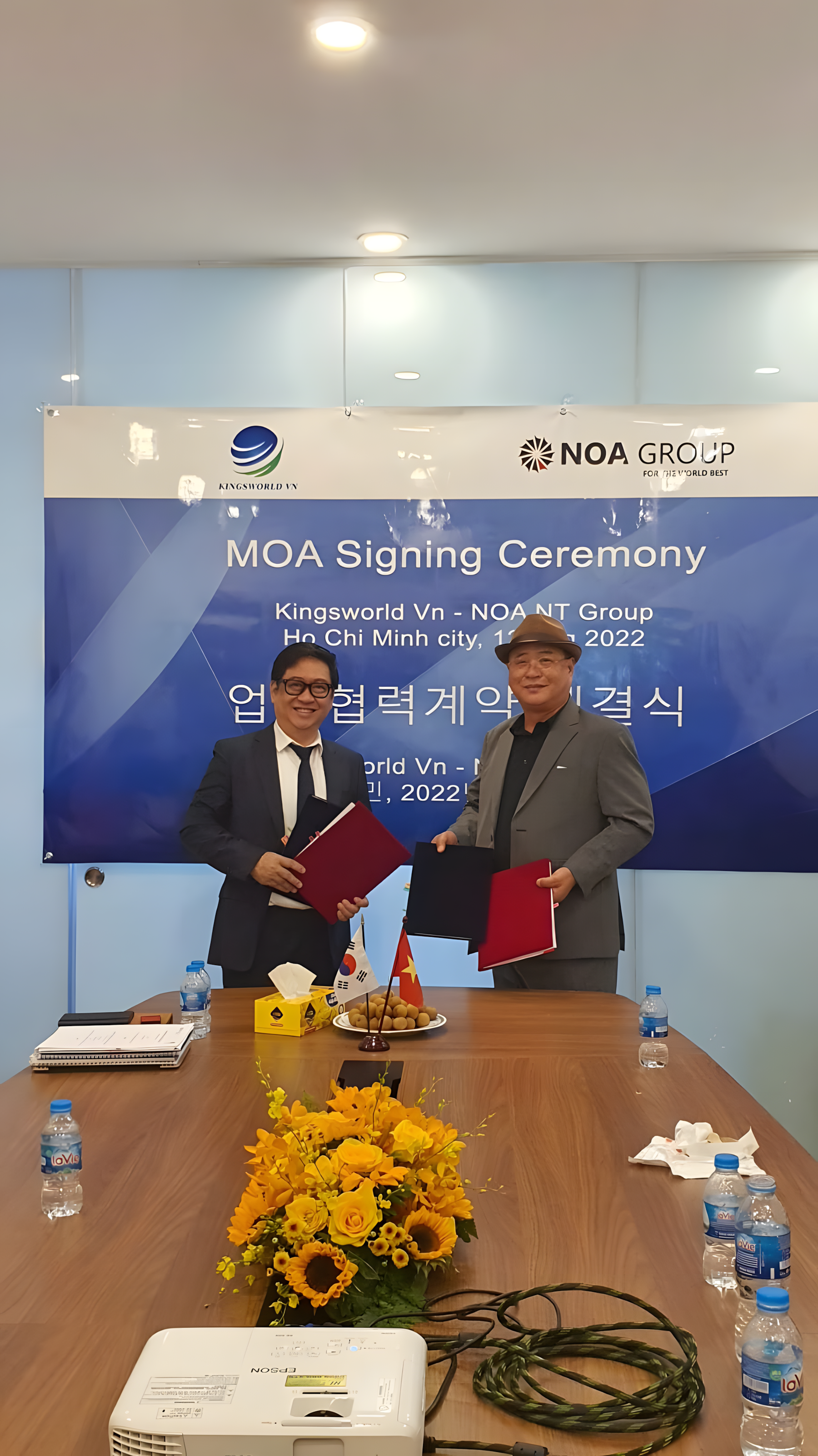 Two men in formal suits shaking hands and holding folders at the MOA signing ceremony, with a large blue banner behind them that reads 'MOA Signing Ceremony' in English and Korean, and a table with flowers, snacks, and water bottles in front.