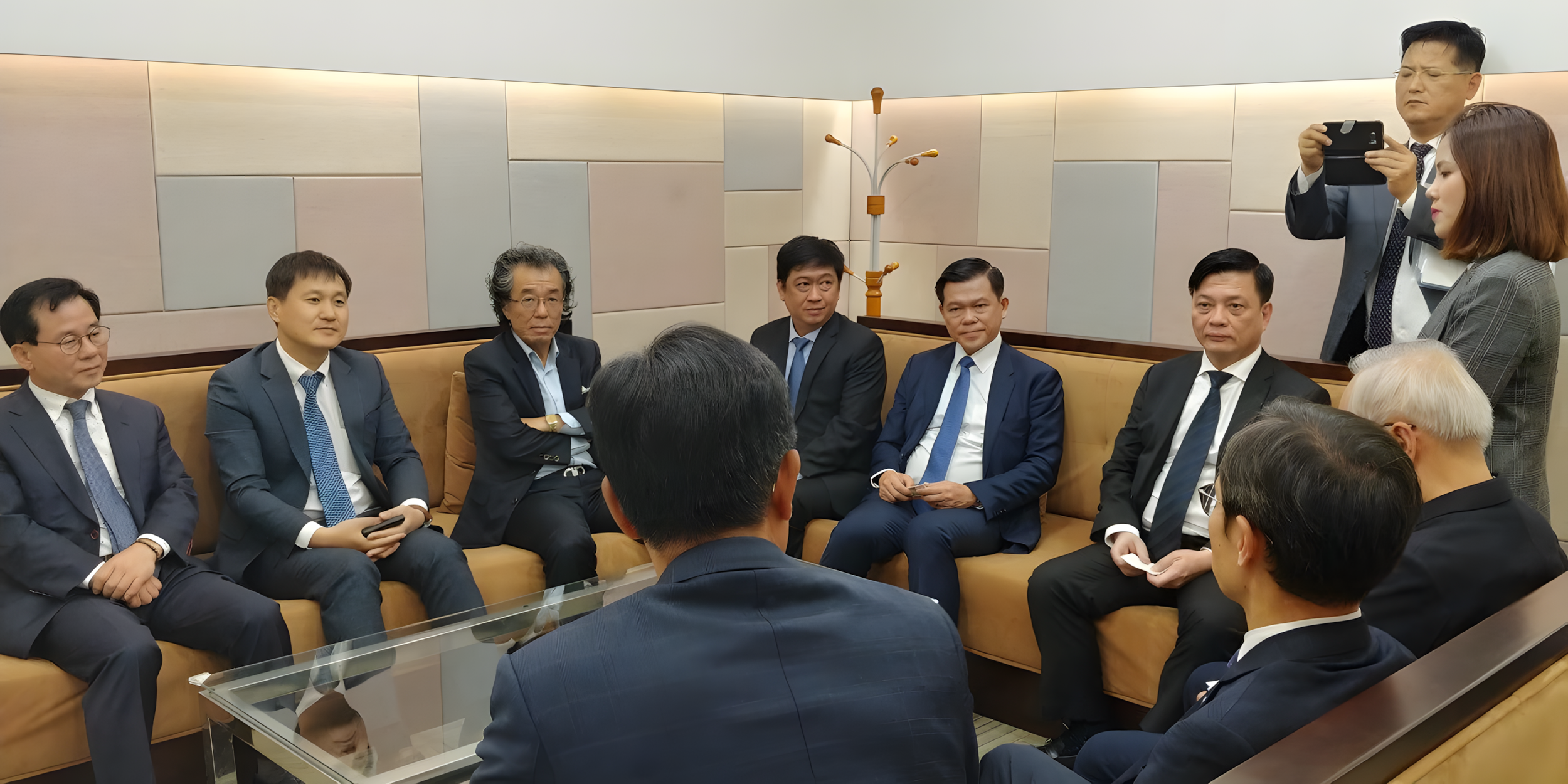 A group of professionally dressed men and women in a meeting room, seated on a sofa and chairs, with some taking notes and others listening attentively. Two individuals are standing, one recording with a phone.