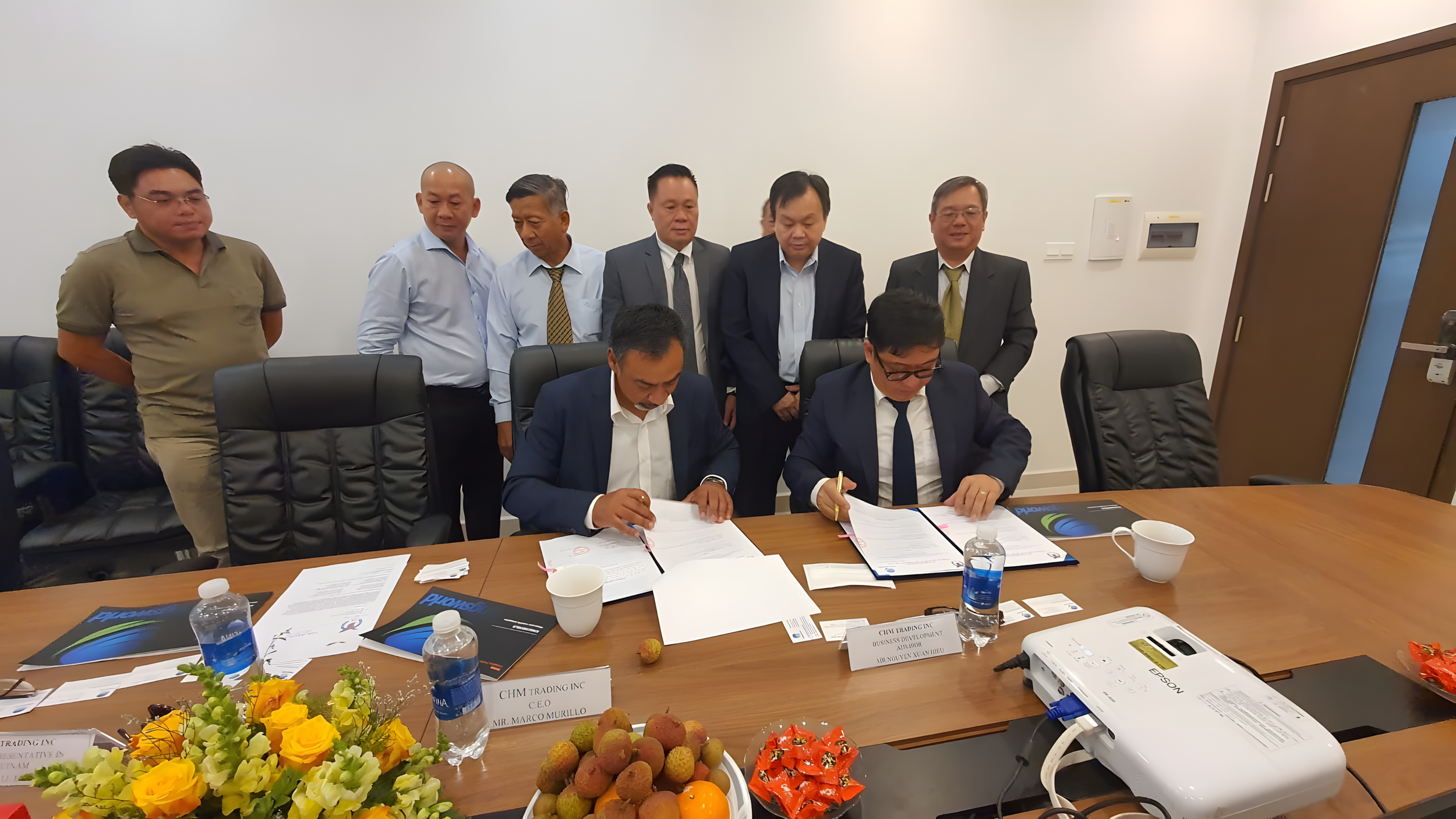 Two men are seated at a conference table signing documents, while eight other men stand behind them observing. The table has bottled water, documents, and a floral arrangement with yellow and white flowers and lychee fruits.