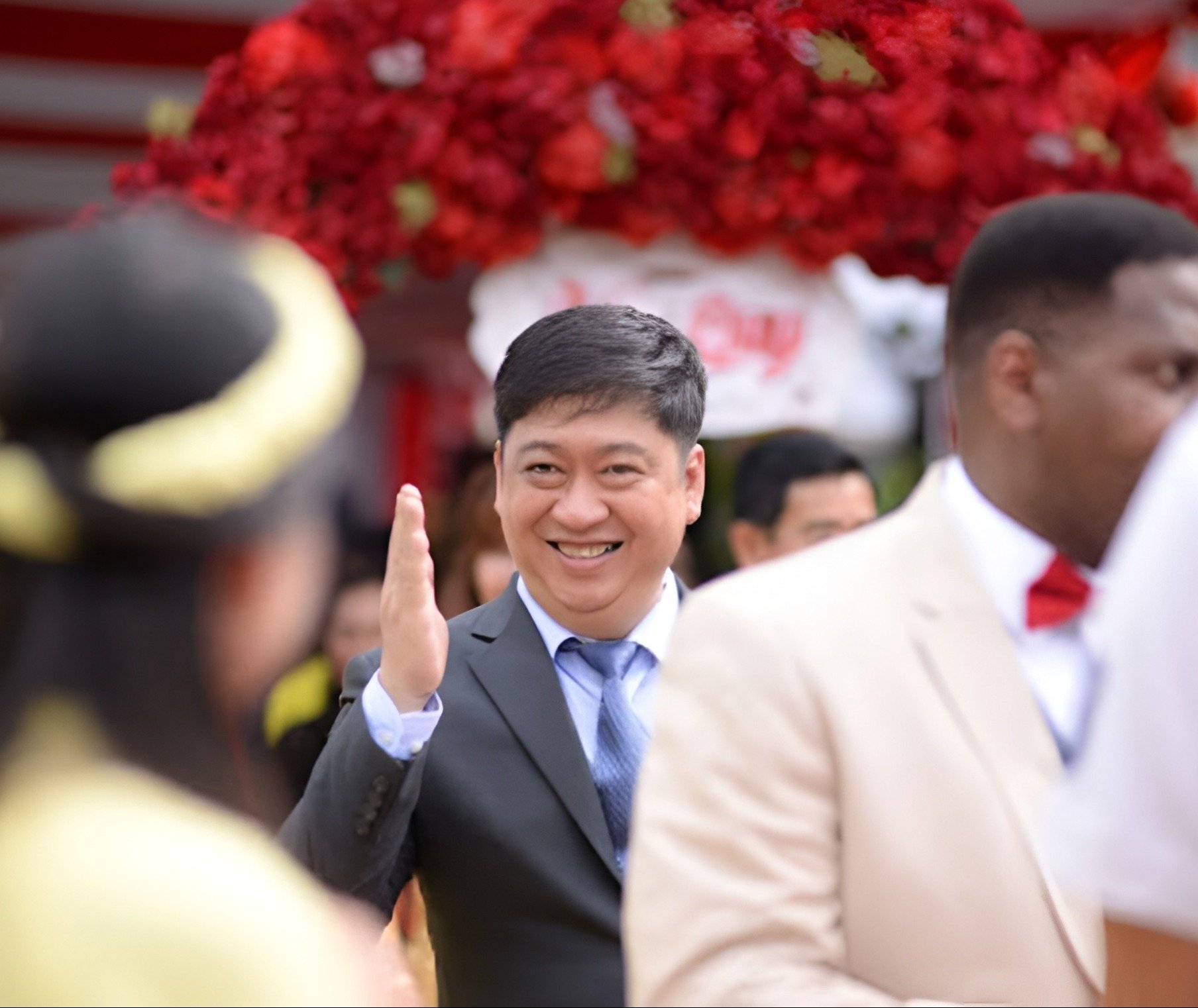 A man waving and smiling, dressed in a dark suit, surrounded by other formally dressed people at an outdoor event with a large red floral decoration in the background.