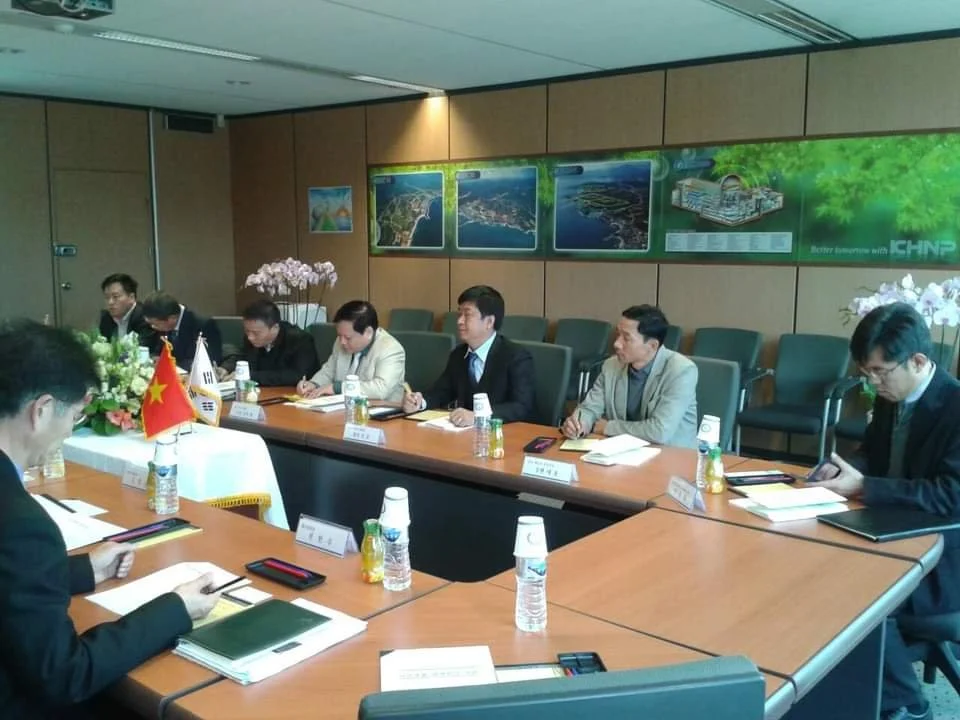 Business meeting with eight people in suits sitting at a U-shaped conference table with notebooks, bottled drinks, and nameplates, in a room decorated with maps and pink flowers.