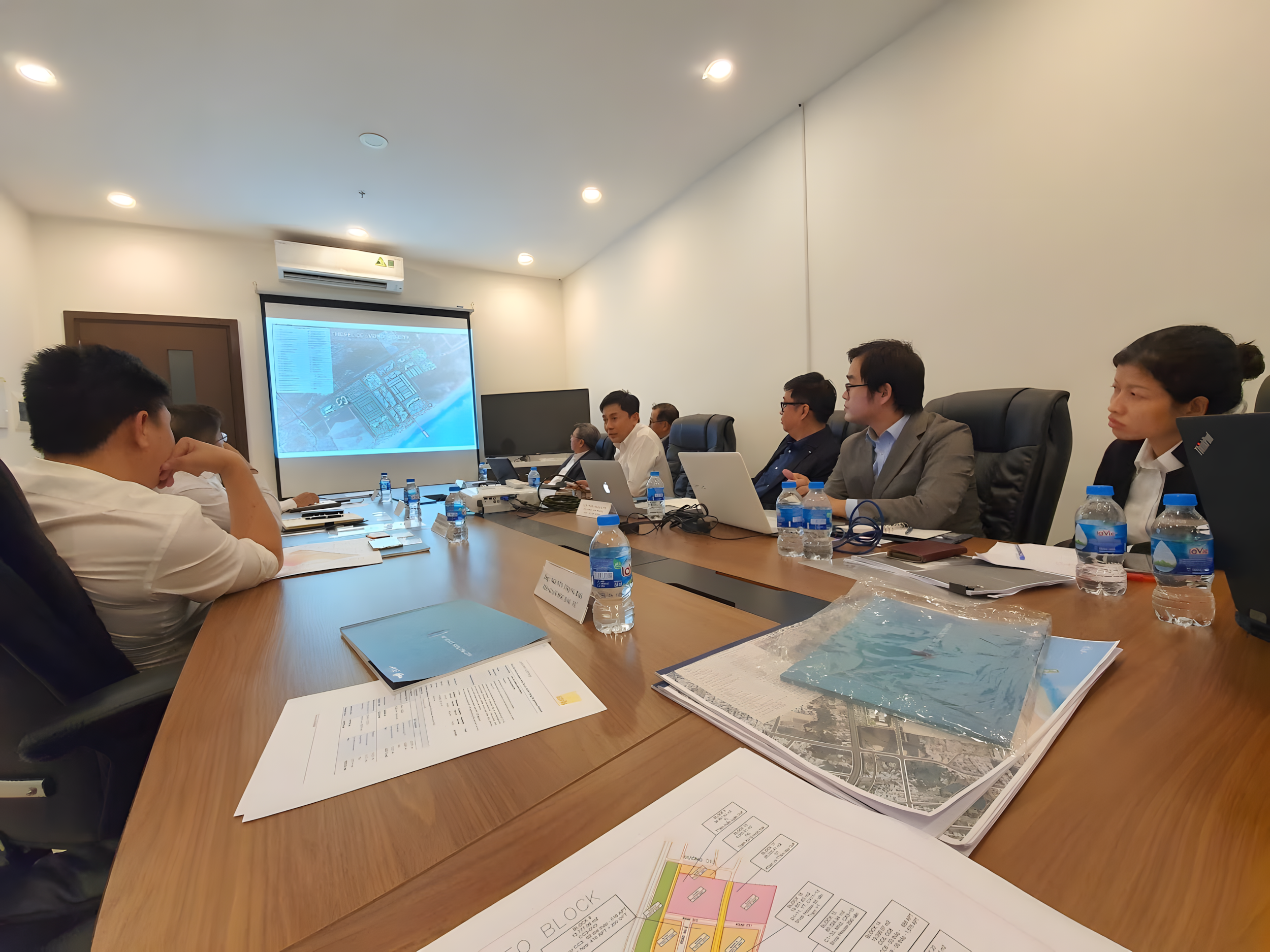 Business meeting with multiple people seated at a conference table, watching a presentation projected on a screen, with documents and bottled water on the table.