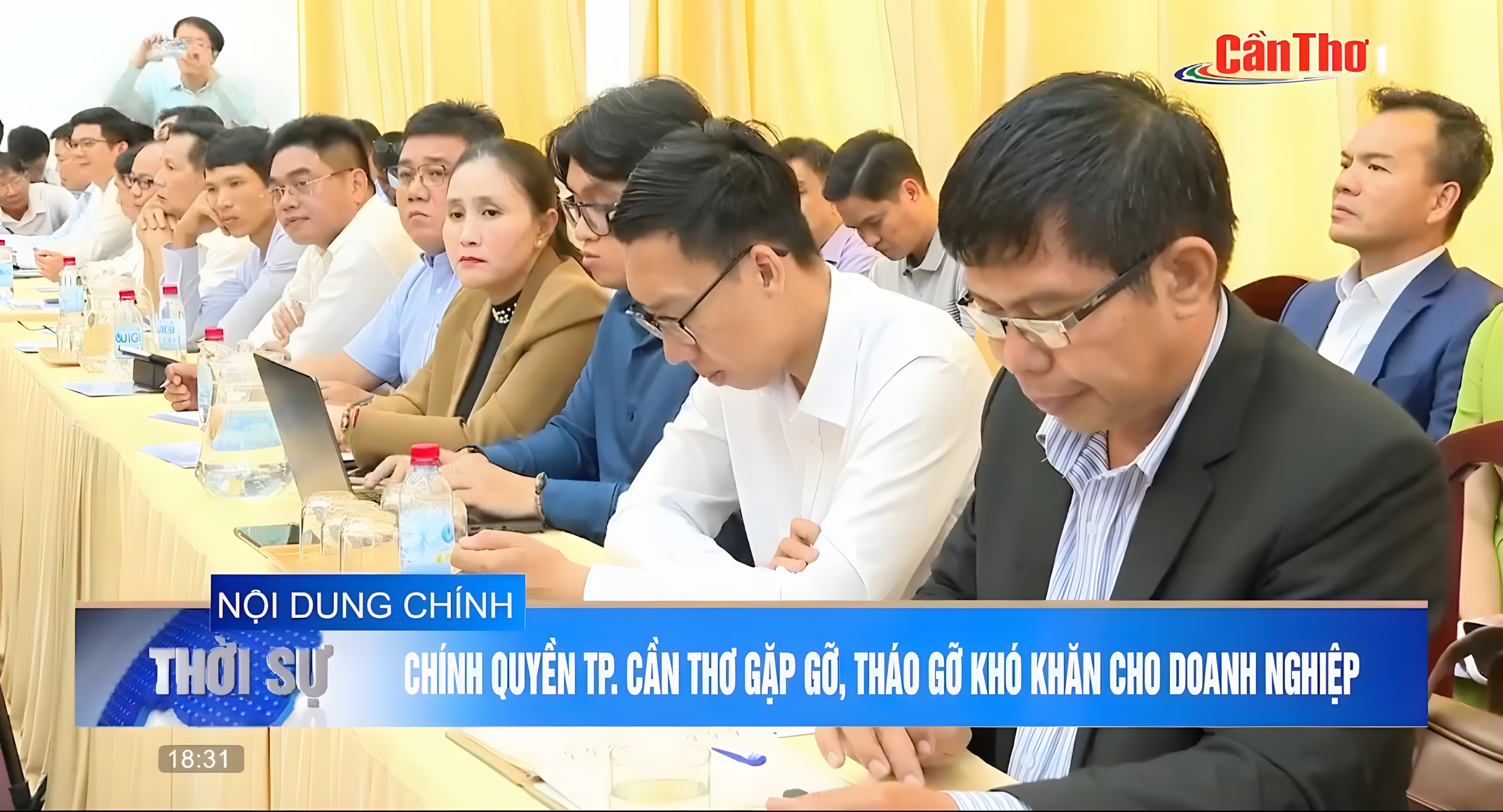 A group of people attending a conference or meeting, sitting at a long table with bottled water and papers, some looking at their phones or listening, in a room with yellow curtains and a person taking a photo in the background.
