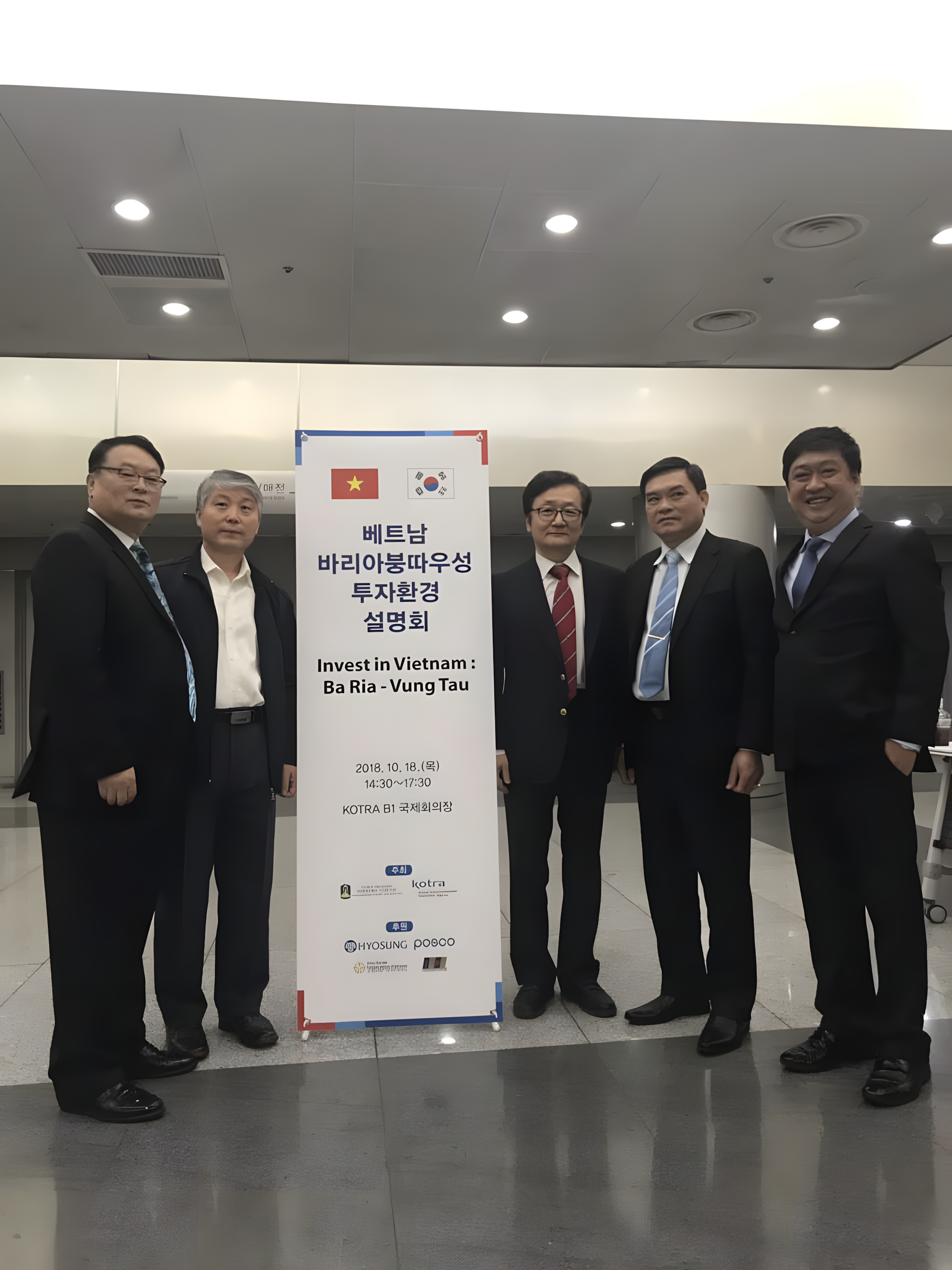 Group of five men in suits standing near a sign about Vietnam investment, at an indoor event, with a plain ceiling and bright lighting.
