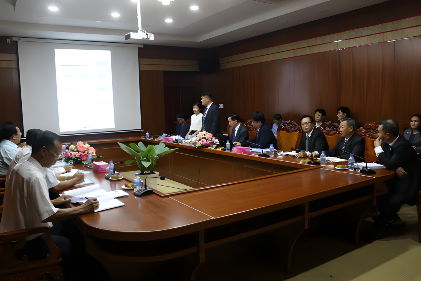 A formal business meeting with multiple individuals seated around a large wooden conference table, some taking notes, with a presenter standing near a projection screen showing a blurred presentation in a wood-paneled conference room.
