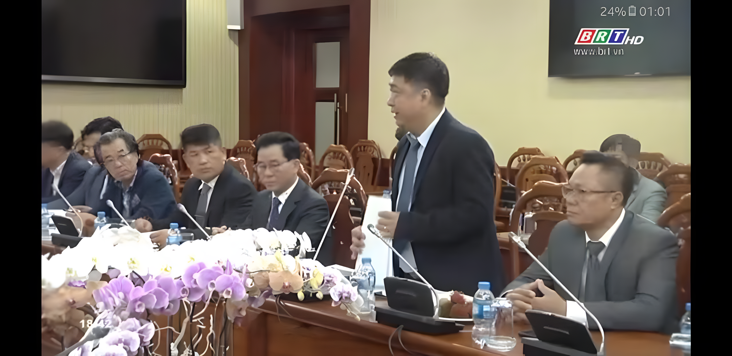 A man in a black suit is speaking at a formal meeting, standing near a long wooden conference table. Several other men in suits are seated around the table, listening. The table has microphones, water bottles, and flowers. A TV screen is on the wall showing a logo and time.
