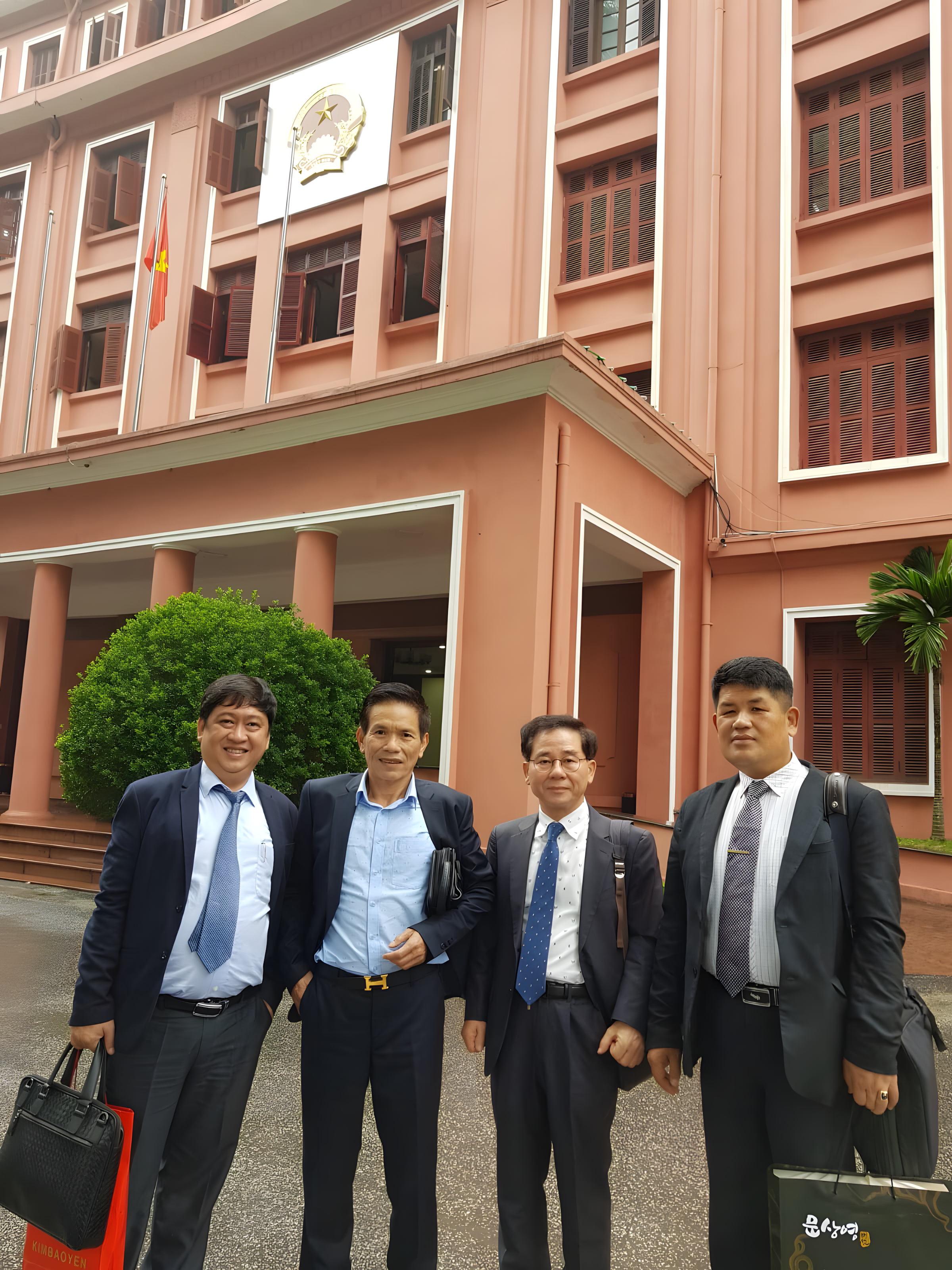 Four men in business suits standing outside a government building with flags and a crest, smiling at the camera.