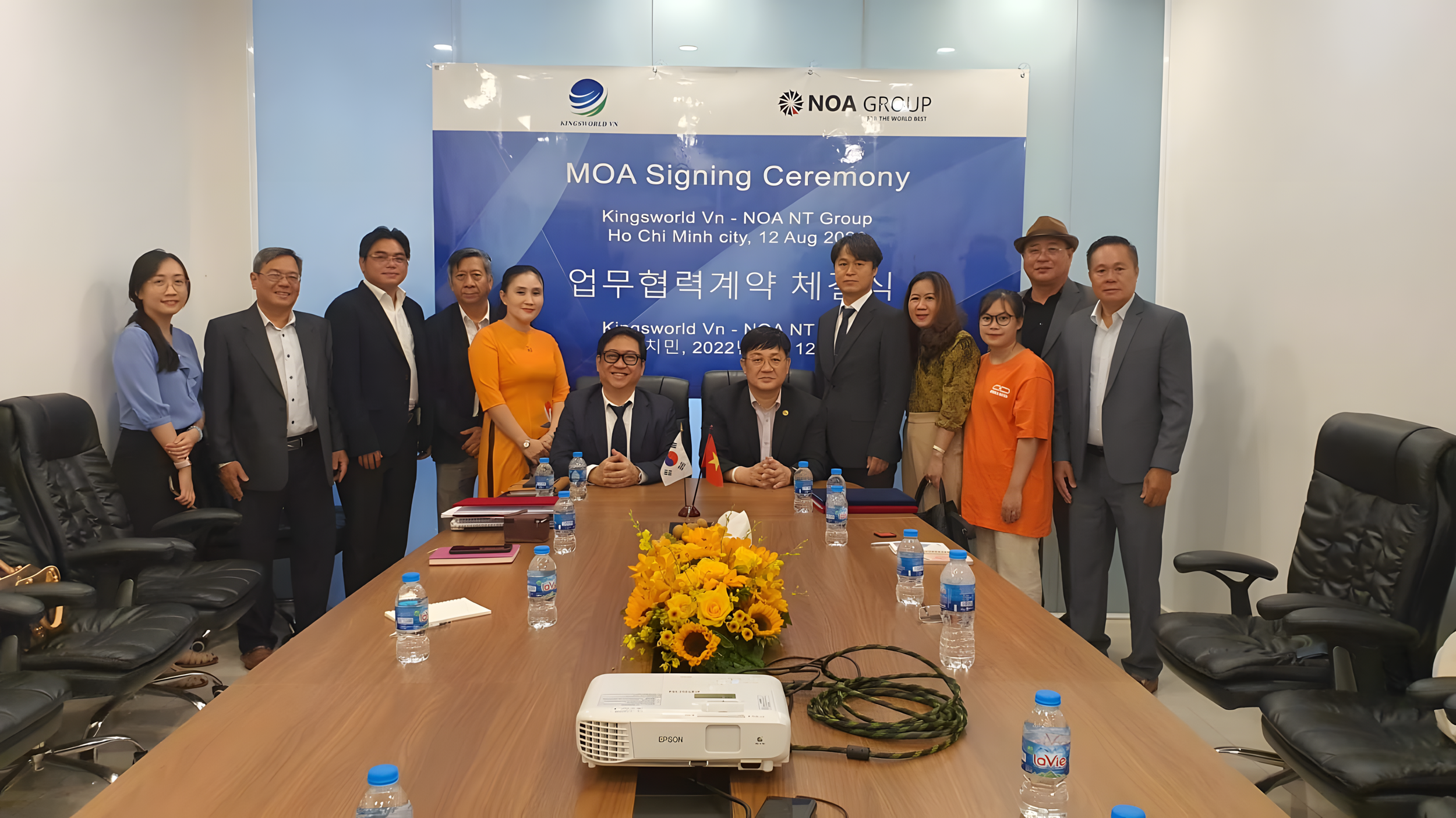 Group of people at a signing ceremony in a conference room, with a table, bottled water, flags, and a blue backdrop.