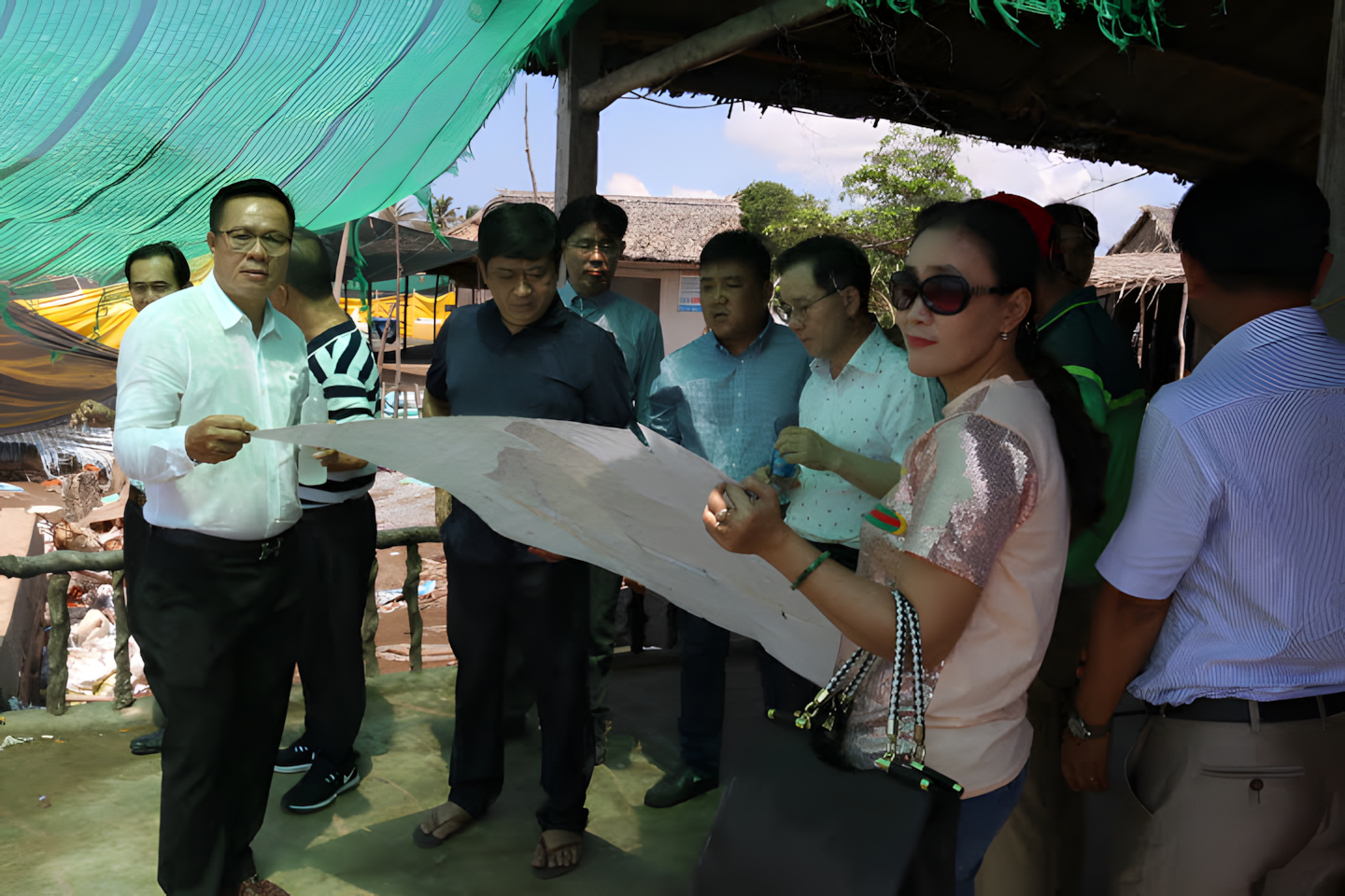 Group of people looking at a large map or blueprint under a makeshift shelter outdoors in a rural area.
