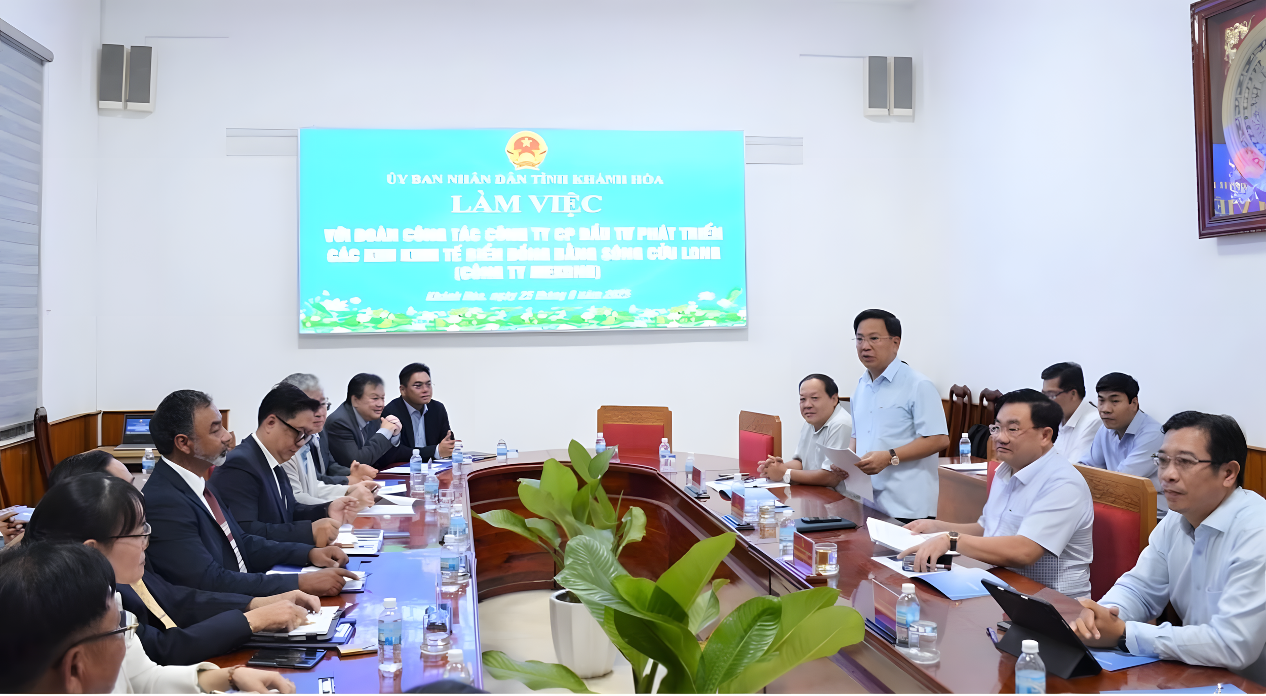 A group of officials in formal attire seated at a conference table in a meeting room, with a man standing and speaking, and a large presentation screen behind them displaying text in Vietnamese.