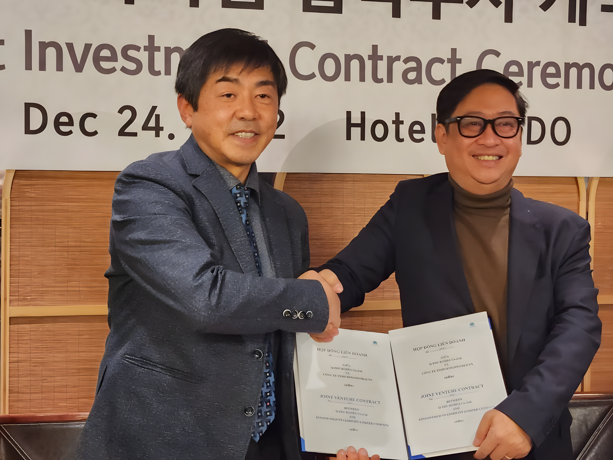 Two men in suits shaking hands and holding a document at a signing ceremony.