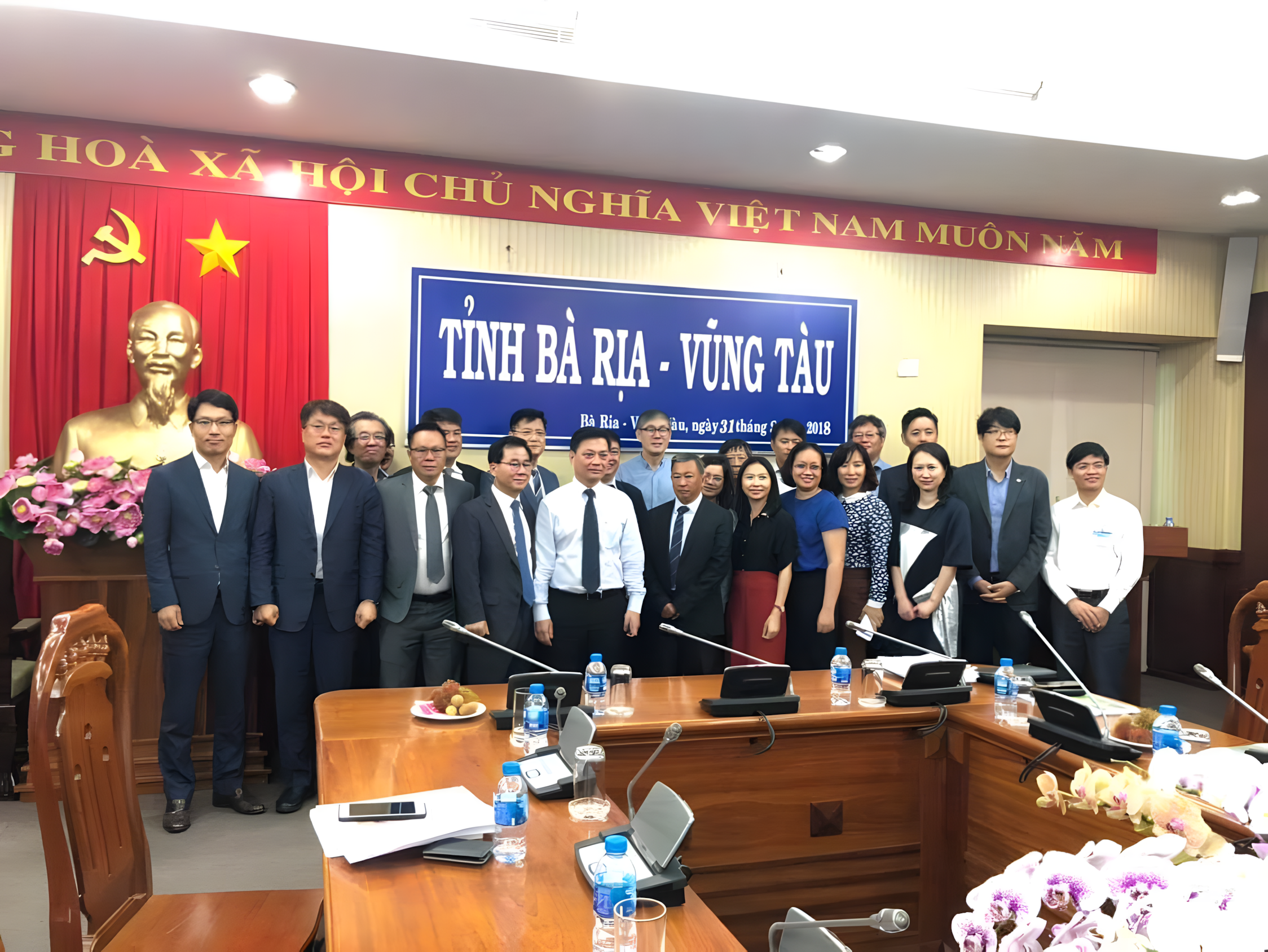 Group of people posing together in a conference room with a blue banner and a bust of Ho Chi Minh in the background, in Vietnam.