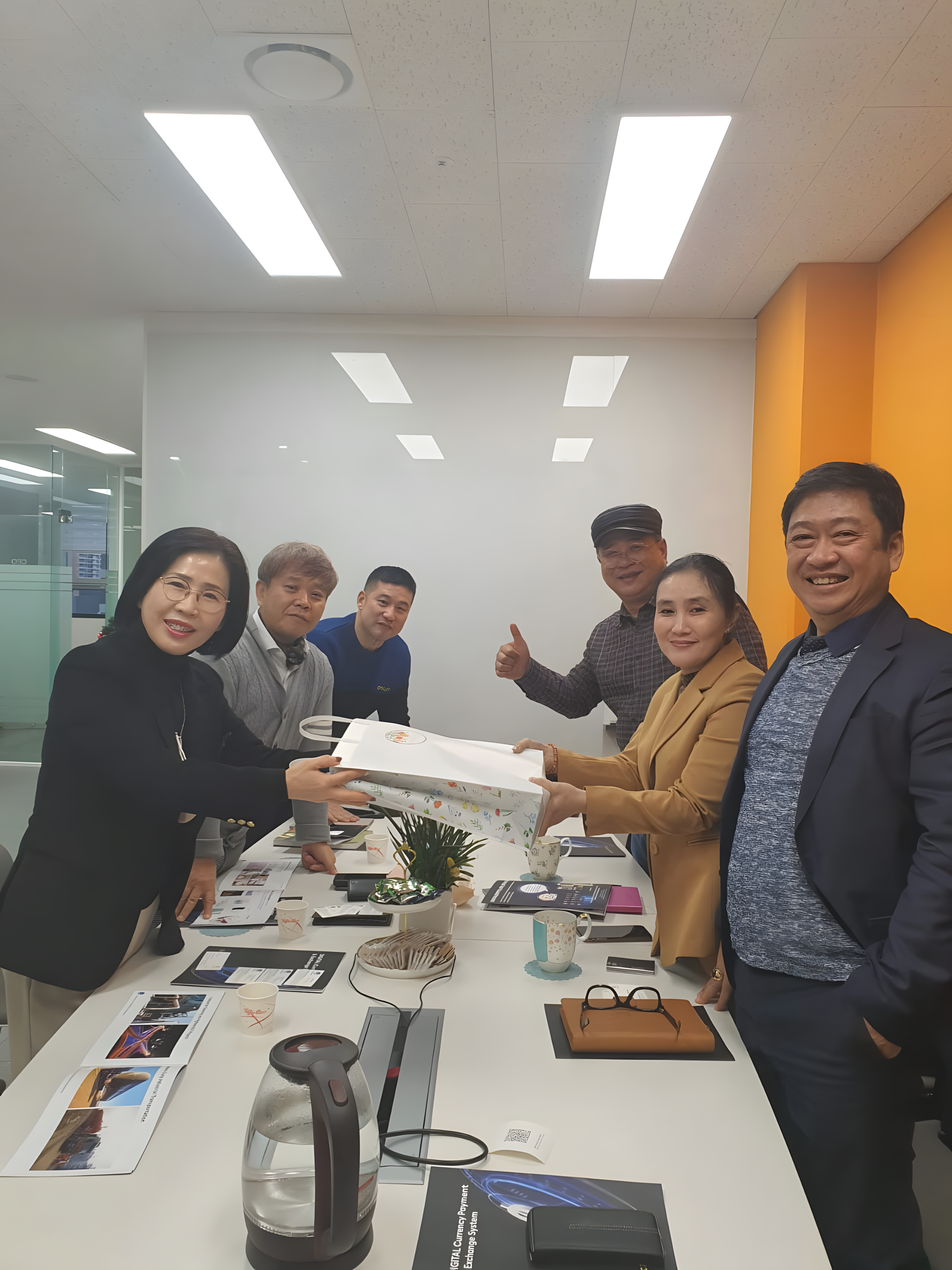 Six people are gathered around a table in a meeting room, exchanging a large gift bag. They are smiling, with one person giving a thumbs up, and there are documents, cups, a water kettle, and a potted plant on the table.