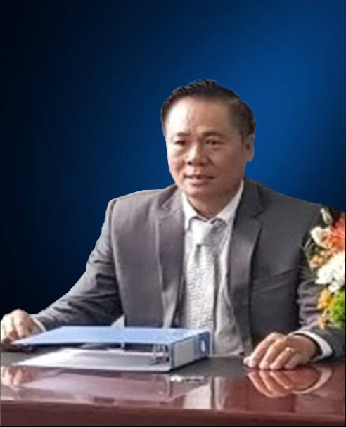 A man in a gray suit sitting at a desk with a blue background, with a binder and a flower arrangement on the desk.