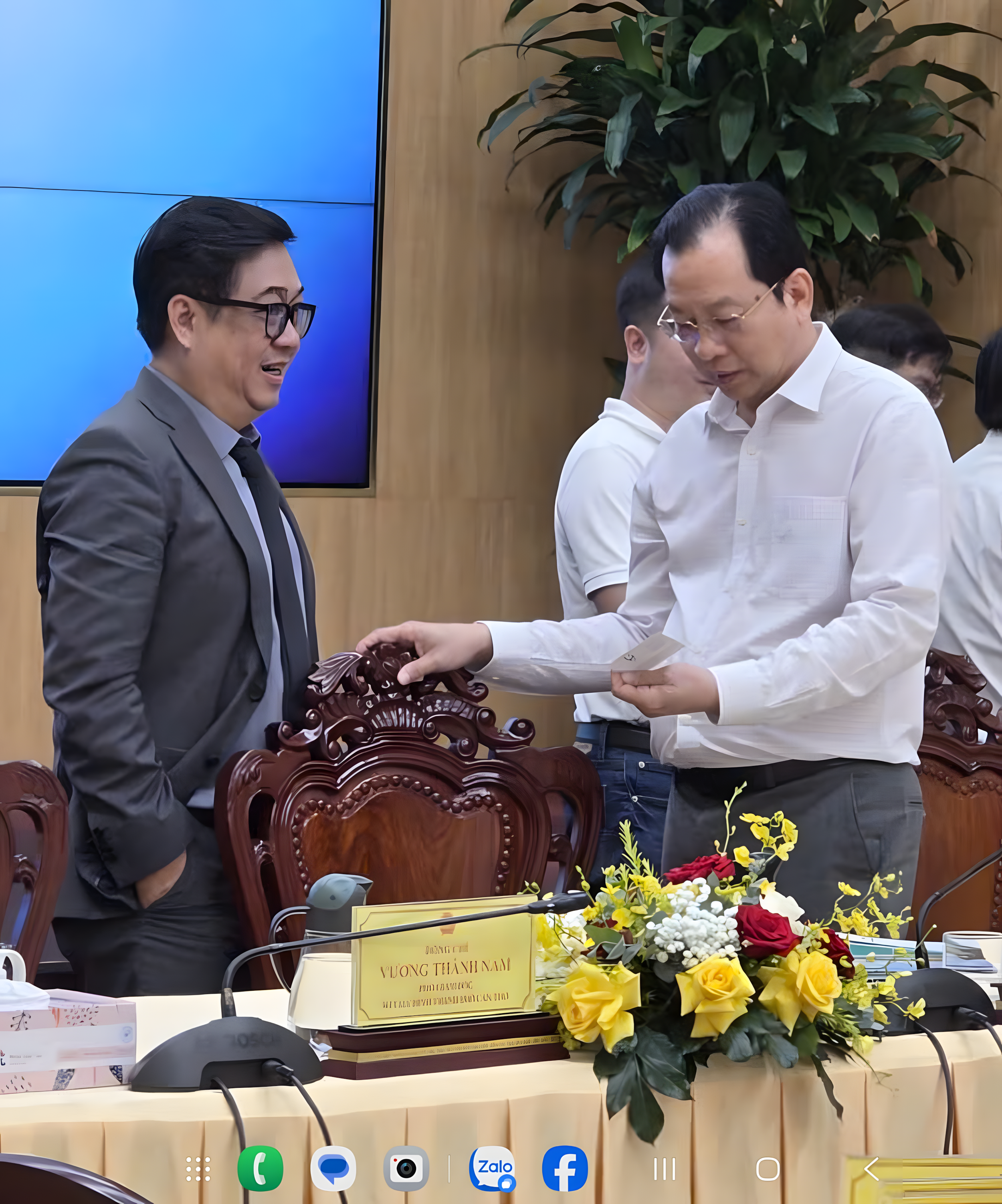 Man in white shirt receiving a document from man in gray suit at a formal meeting with a flower arrangement and nameplate on table.