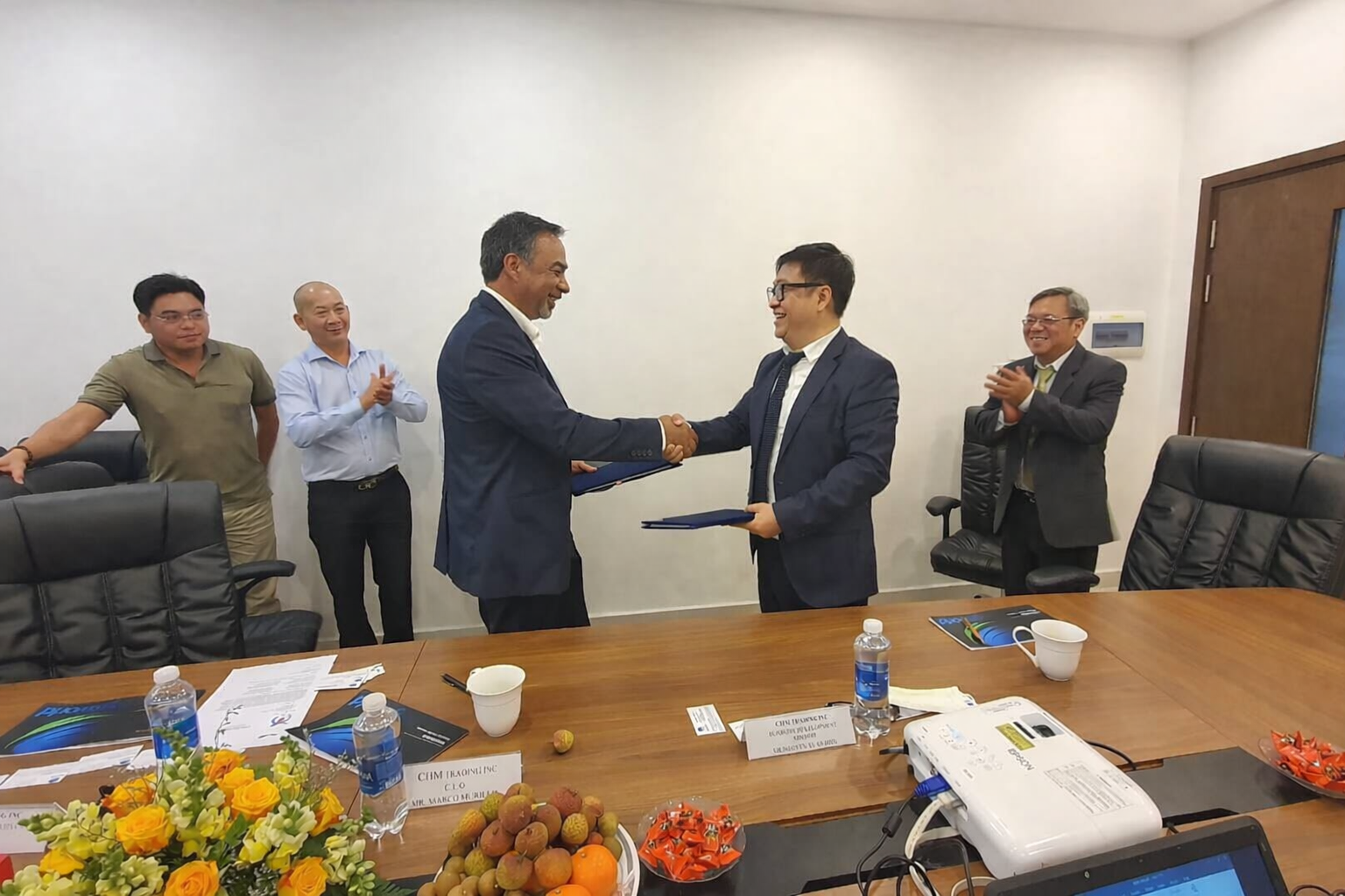 Two men in suits shake hands in a conference room, surrounded by four other people clapping and smiling, with a table of documents, water bottles, fruit, and flowers in the foreground.