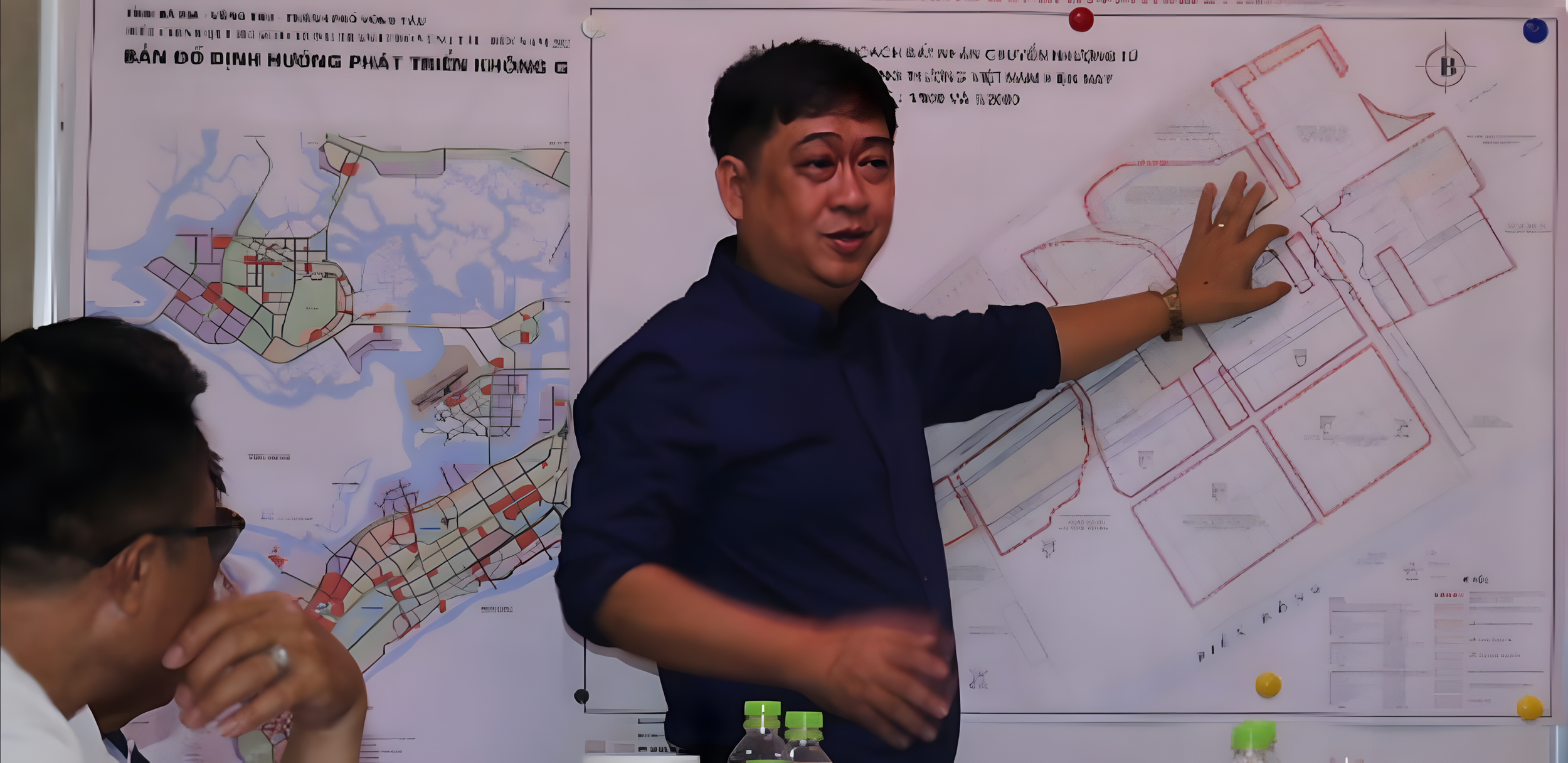 A man standing in front of a large map on a wall, pointing to a section of the map with his right hand, while talking to others and explaining the map. There are two water bottles on a table in front of him, and another person is partially visible in the foreground.