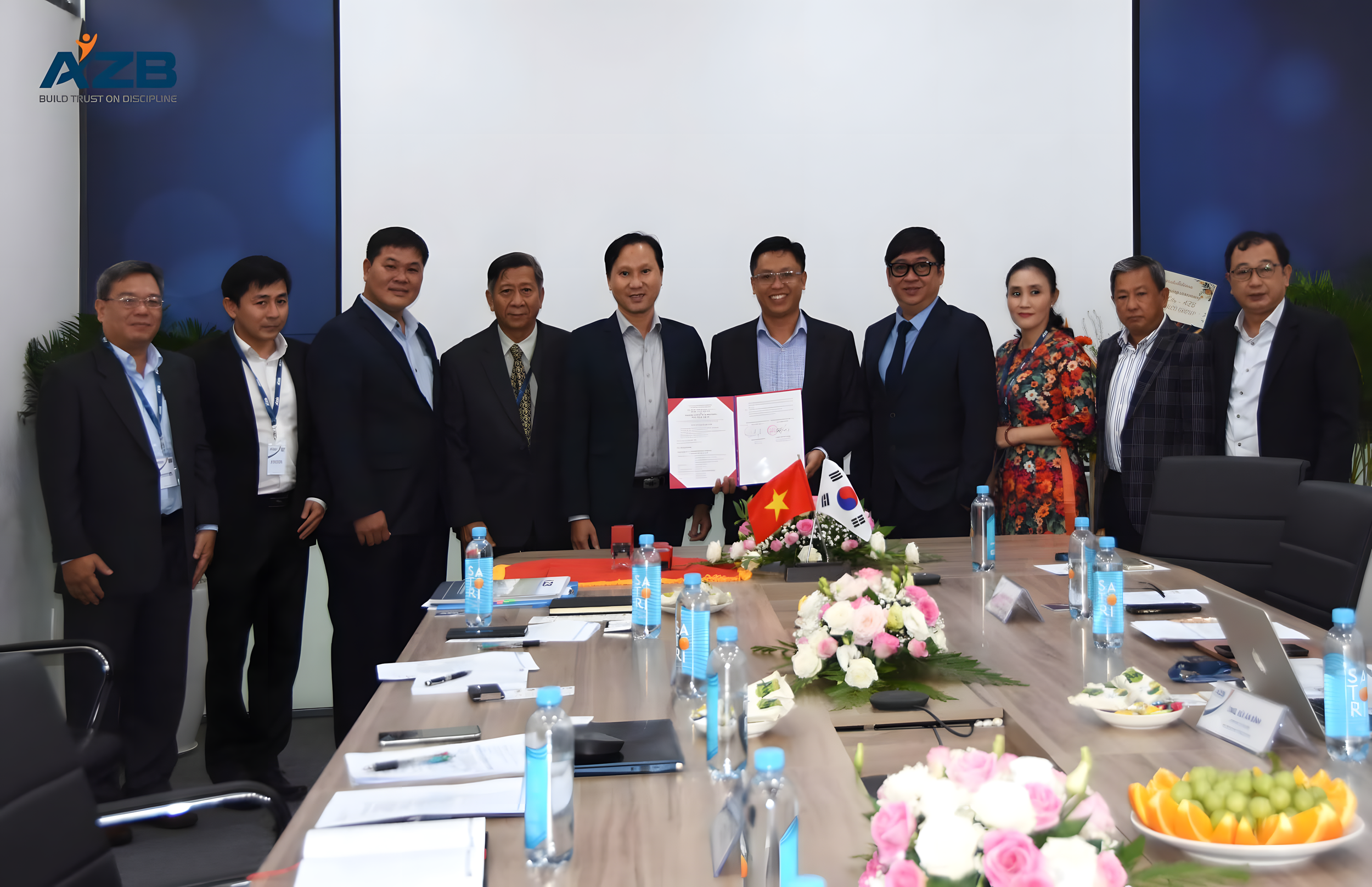 A group of eleven business professionals, including men and women, standing behind a conference table decorated with flowers and flags of Vietnam and South Korea. The individuals are smiling, and two of them are holding a document. The conference roo