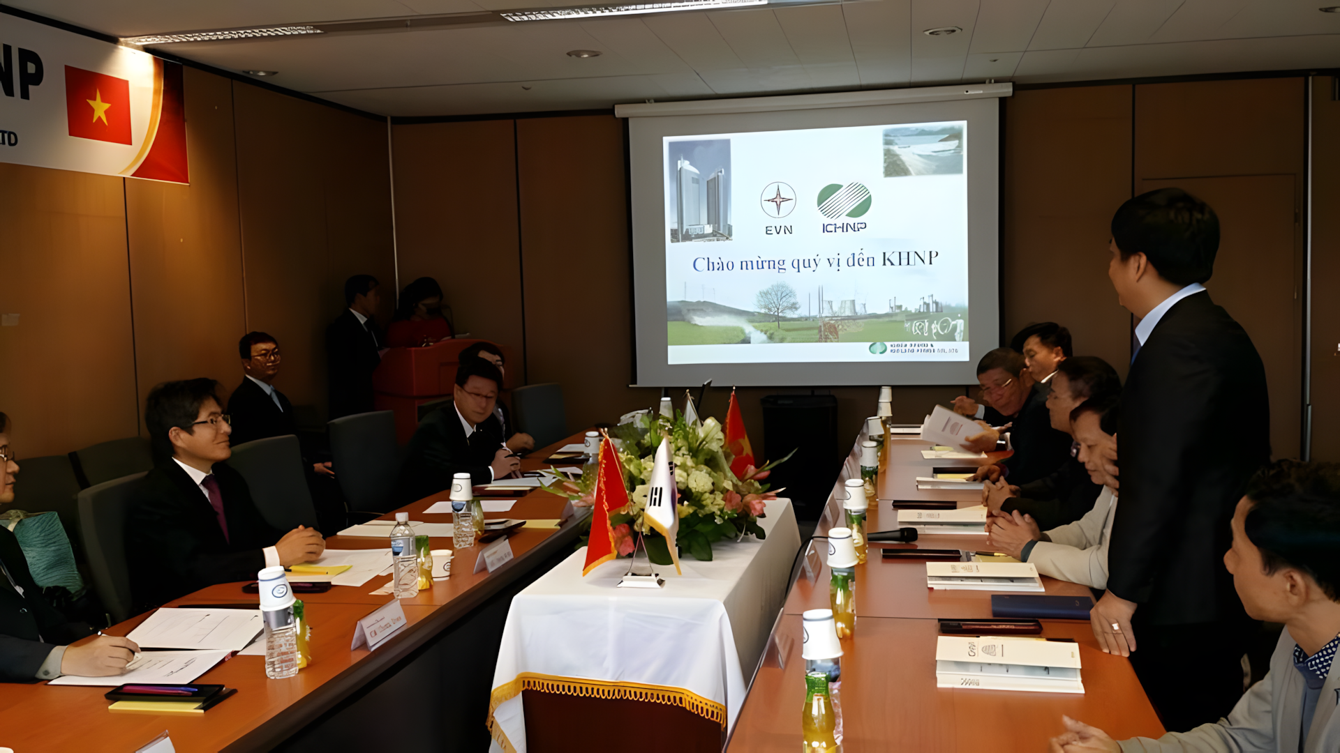A group of business people sitting around a conference table with flags, notebooks, and drinks, attending a meeting or presentation in a well-lit conference room. There is a large screen displaying logos and text in Vietnamese.
