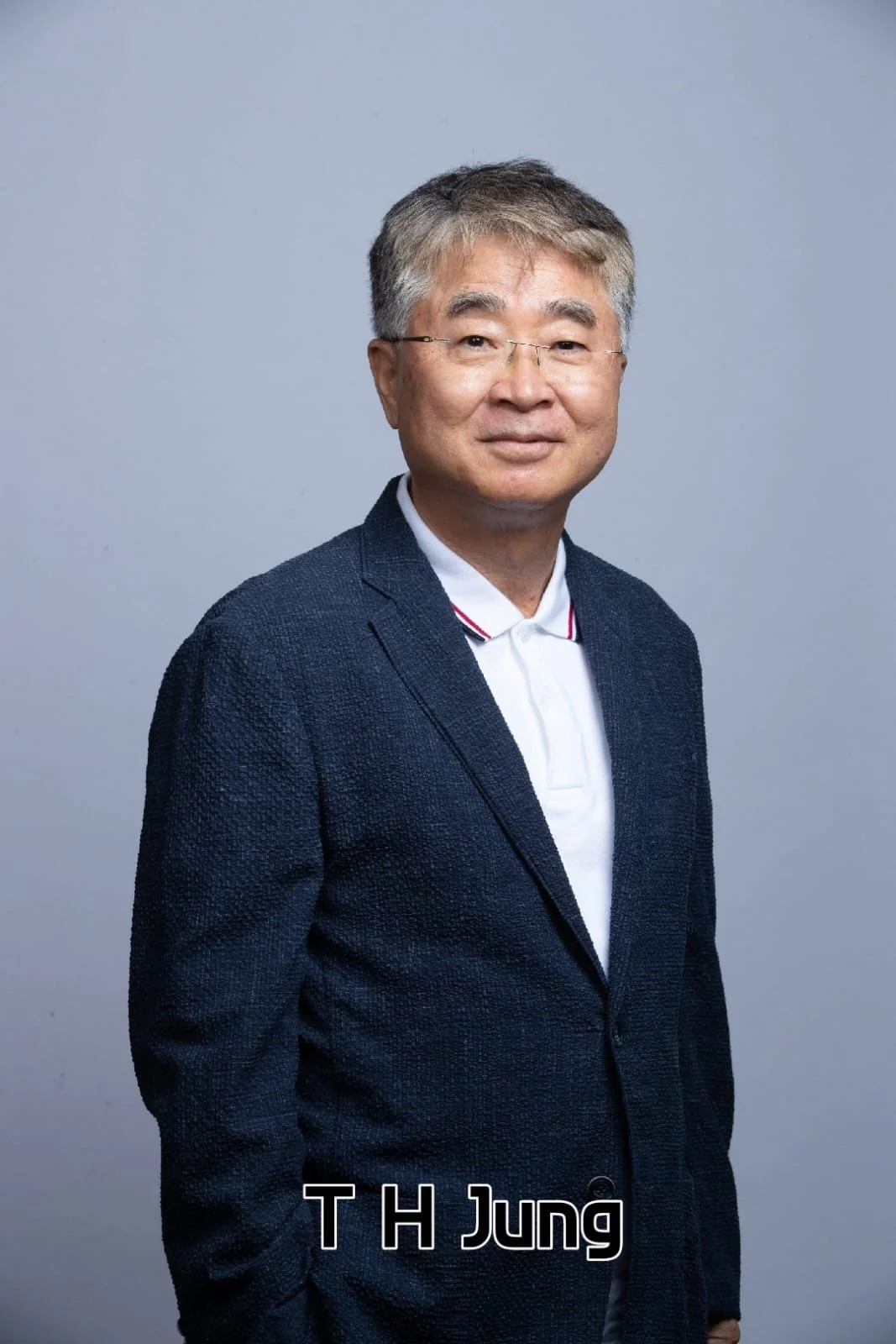 Portrait of T H Jung wearing glasses, a white polo shirt, and a dark blazer, standing against a plain gray background.