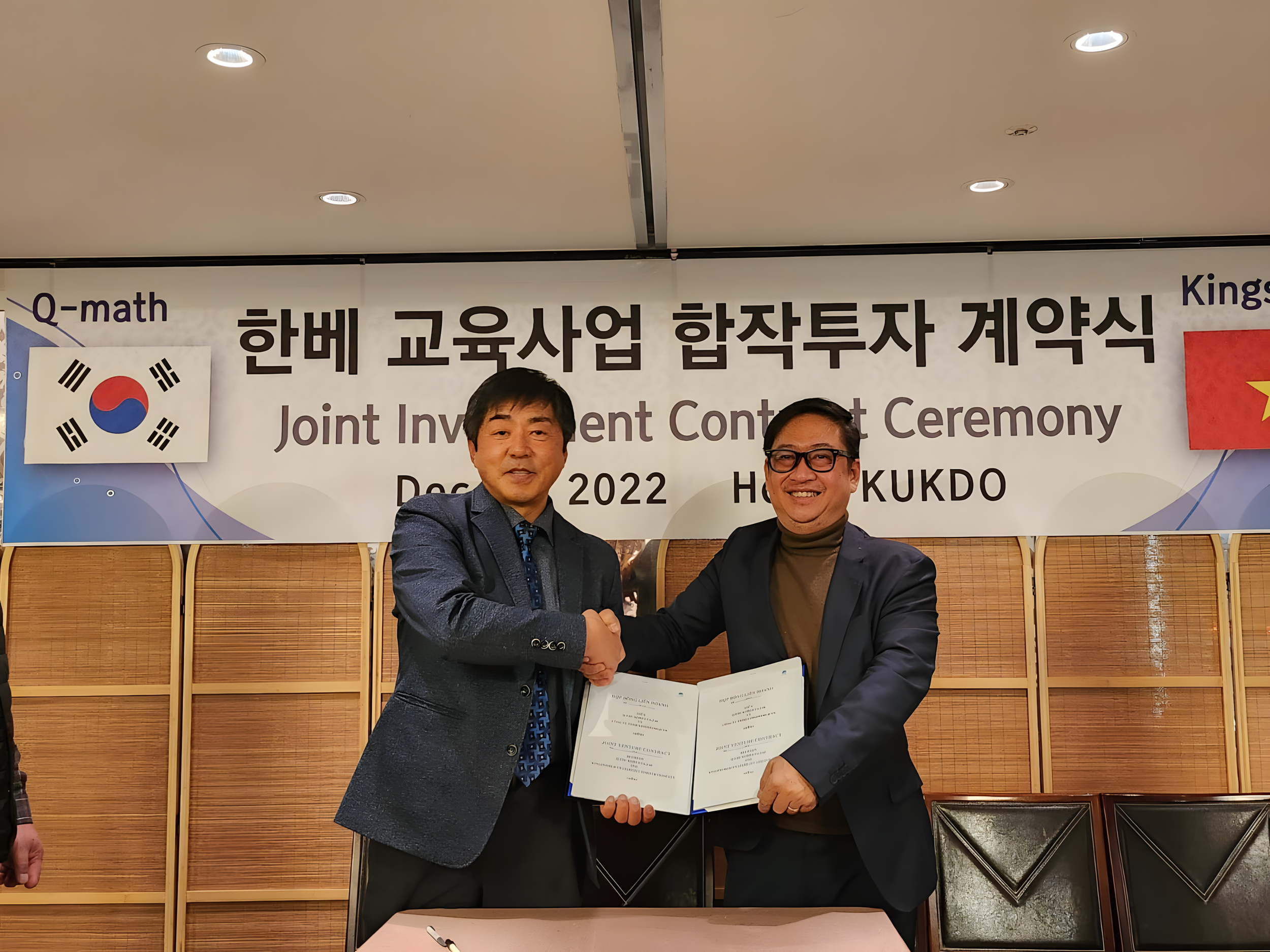 Two men shaking hands, holding a document, at a signing ceremony with a banner in the background. The banner has Korean and English text, flags of South Korea and Vietnam, and the event mentions a joint investment contract.
