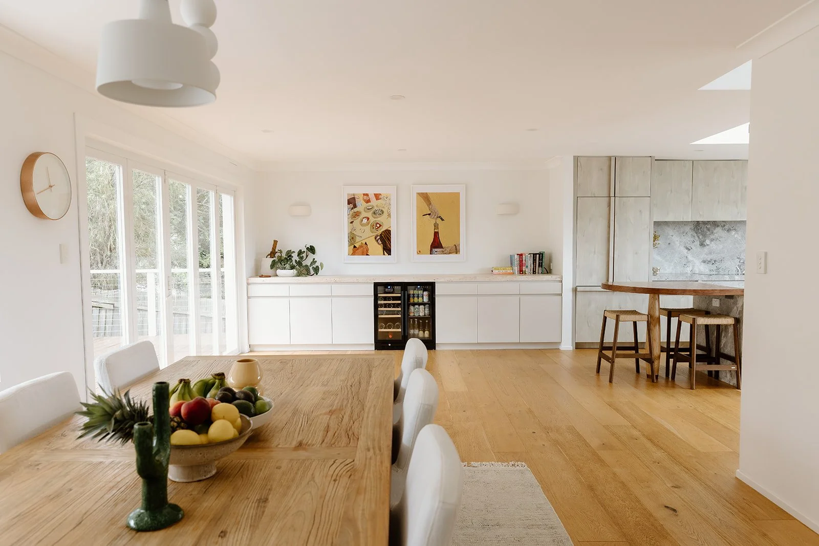 Dining space design Central Coast with timber, travertine stone and soft neutral palette featuring white walls, artwork and a small wine fridge.
