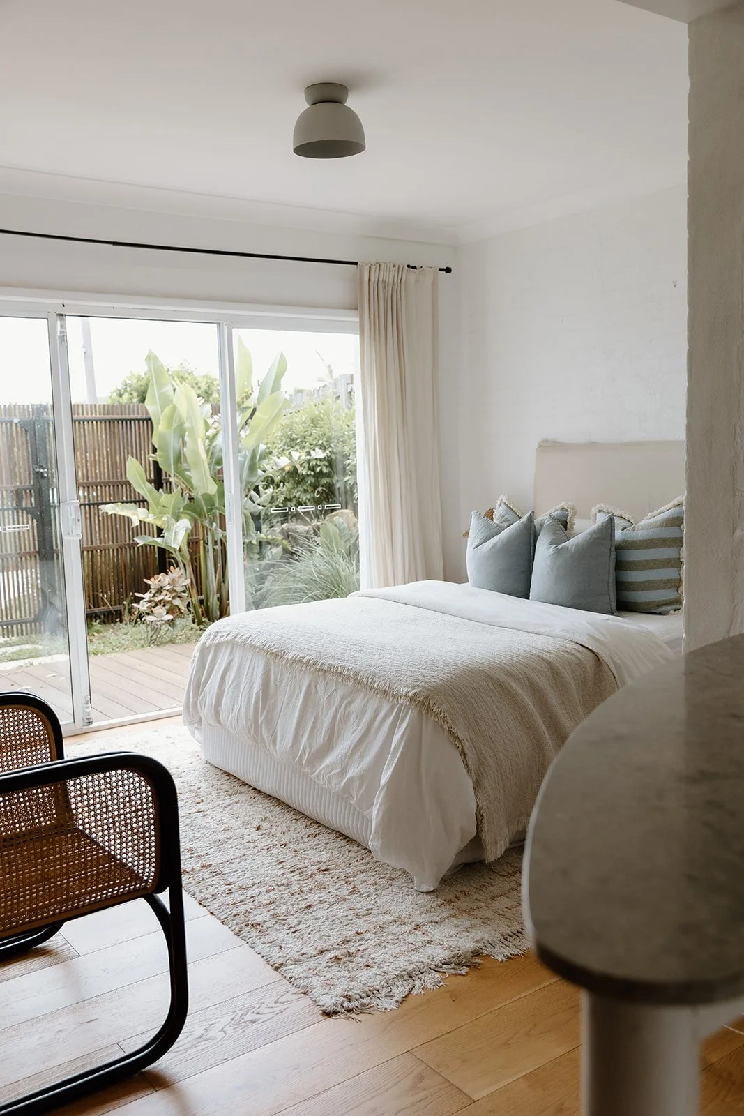 Airbnb studio interior design Central Coast with soft neutral tones and natural materials featuring kitchenette, linen curtains, cane armchair and throw pillows. 