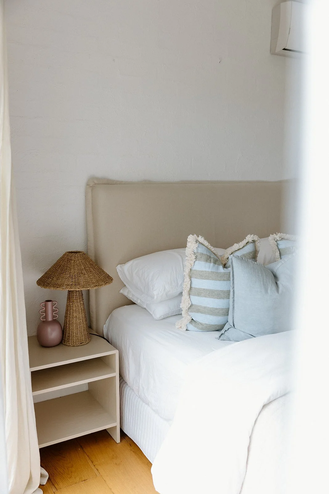 Bedroom design Central Coast with soft tones and layered natural textures featuring light blue and beige striped throw pillows, linen bedhead and a woven lamp.