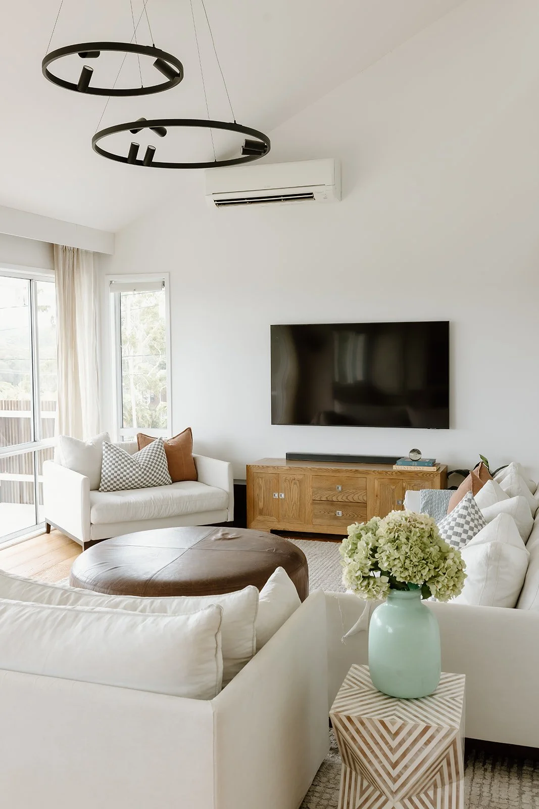 Living room design Central Coast with warm tones and natural materials by Everyday Designs featuring white linen sofas, throw pillows, leather ottoman, striped side table and blue vase. 