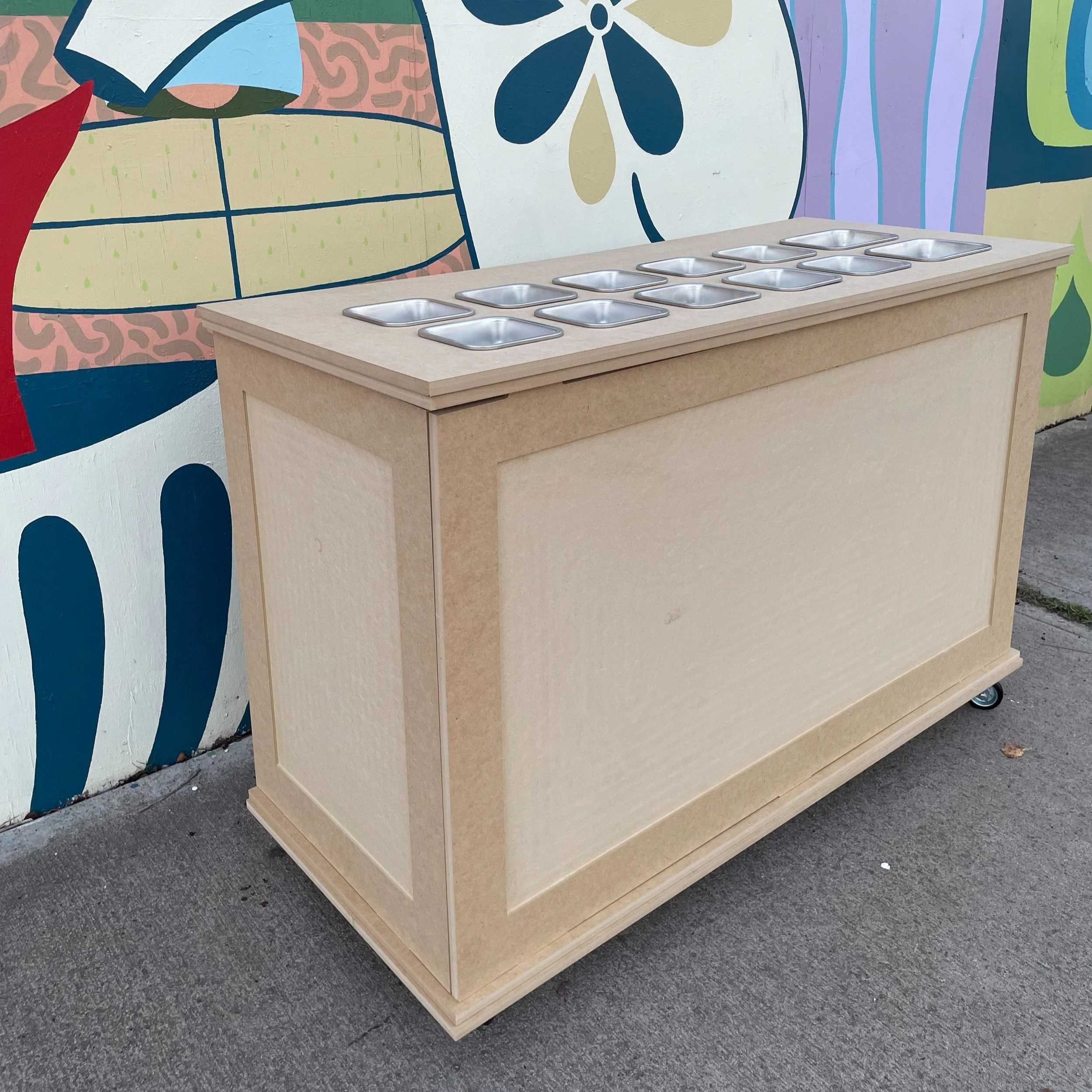A wooden food cart with eight metal compartments on top, set against a colorful mural background on a sidewalk.