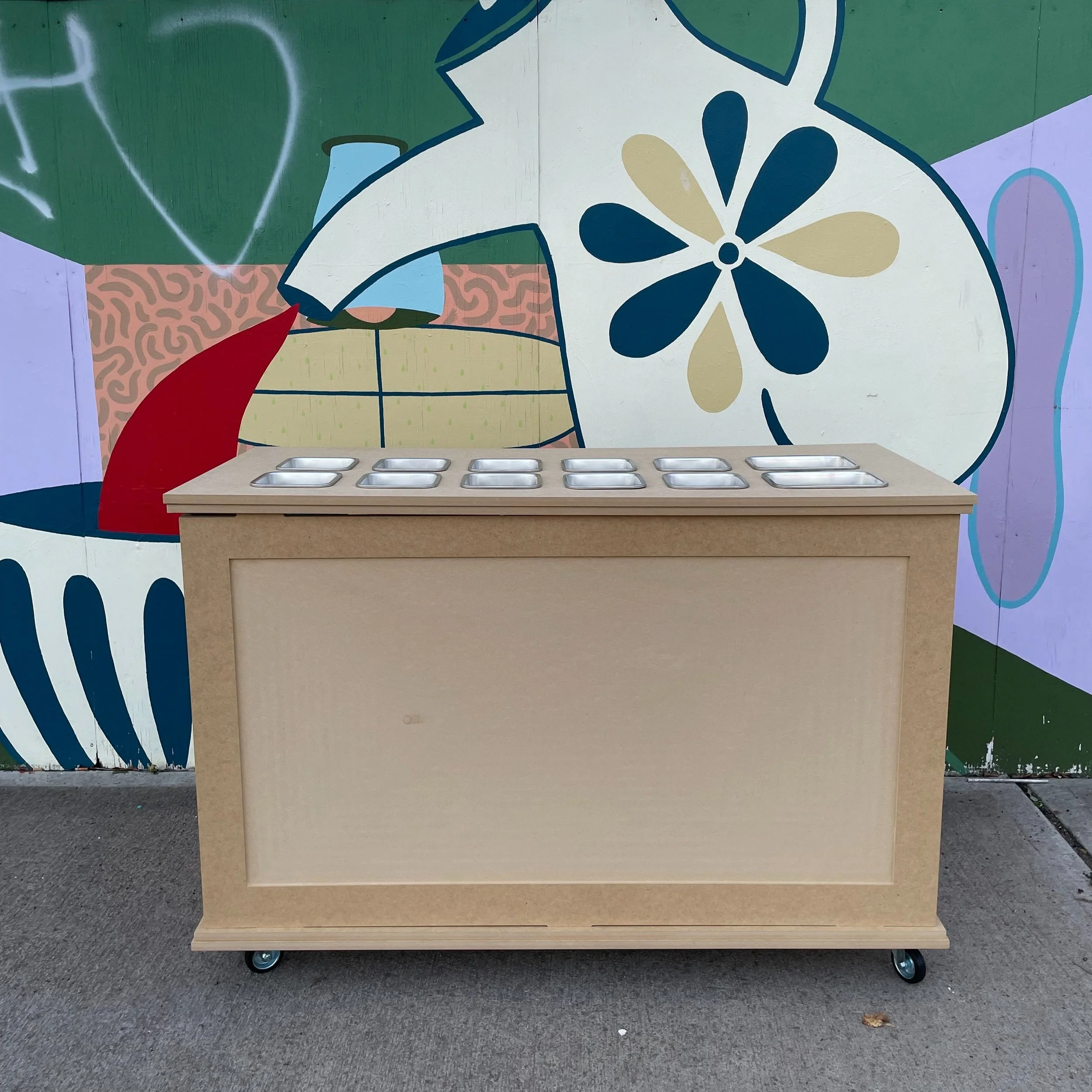 A beige wooden table on wheels with multiple small metal containers on top, placed in front of a colorful, abstract mural on a wall.