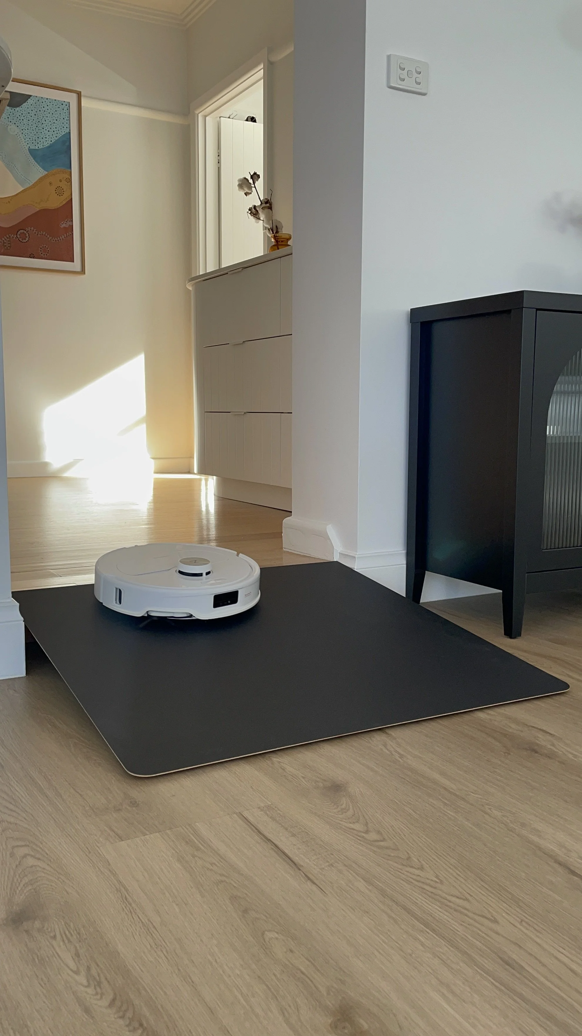 A white robotic vacuum cleaner on a black cleaning mat in a hardwood-floored living room.