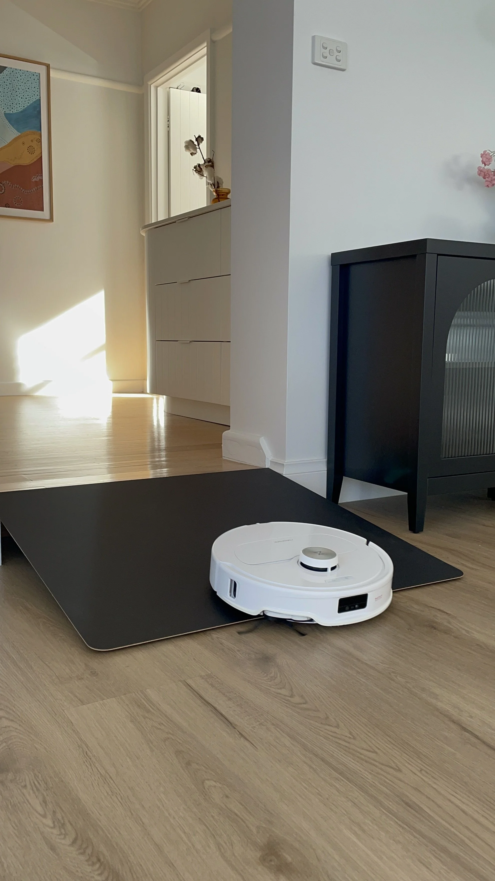 A white robotic vacuum cleaner on a black mat in a home with wooden flooring and white walls.