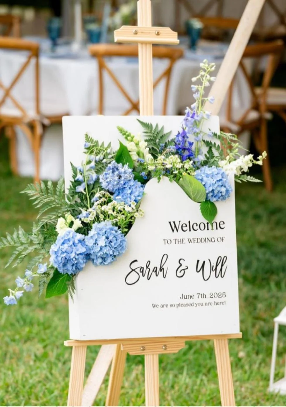 Wedding welcome sign with blue hydrangeas and greenery on an easel, displaying names Sarah and Will, date June 7th, 2025, in an outdoor setting with tables and chairs in the background.