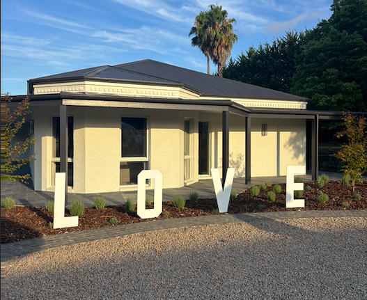 A single-story house with a dark roof and beige walls, decorated with large white letters spelling out 'LOVE' in the front yard during daytime.