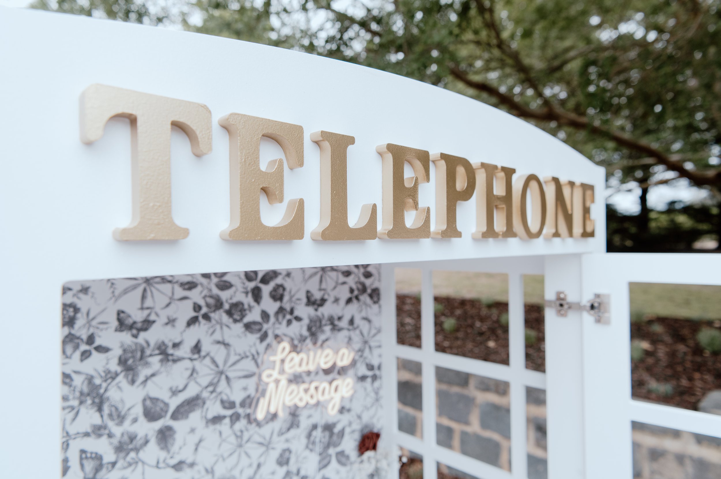 White collapsible telephone booth purchase with vintage rotary phone, floral wallpaper, brass handle, mirrored ceiling, tiled base, and 'Leave a Message' LED sign—perfect for weddings, parties, and audio guestbooks in Melbourne.