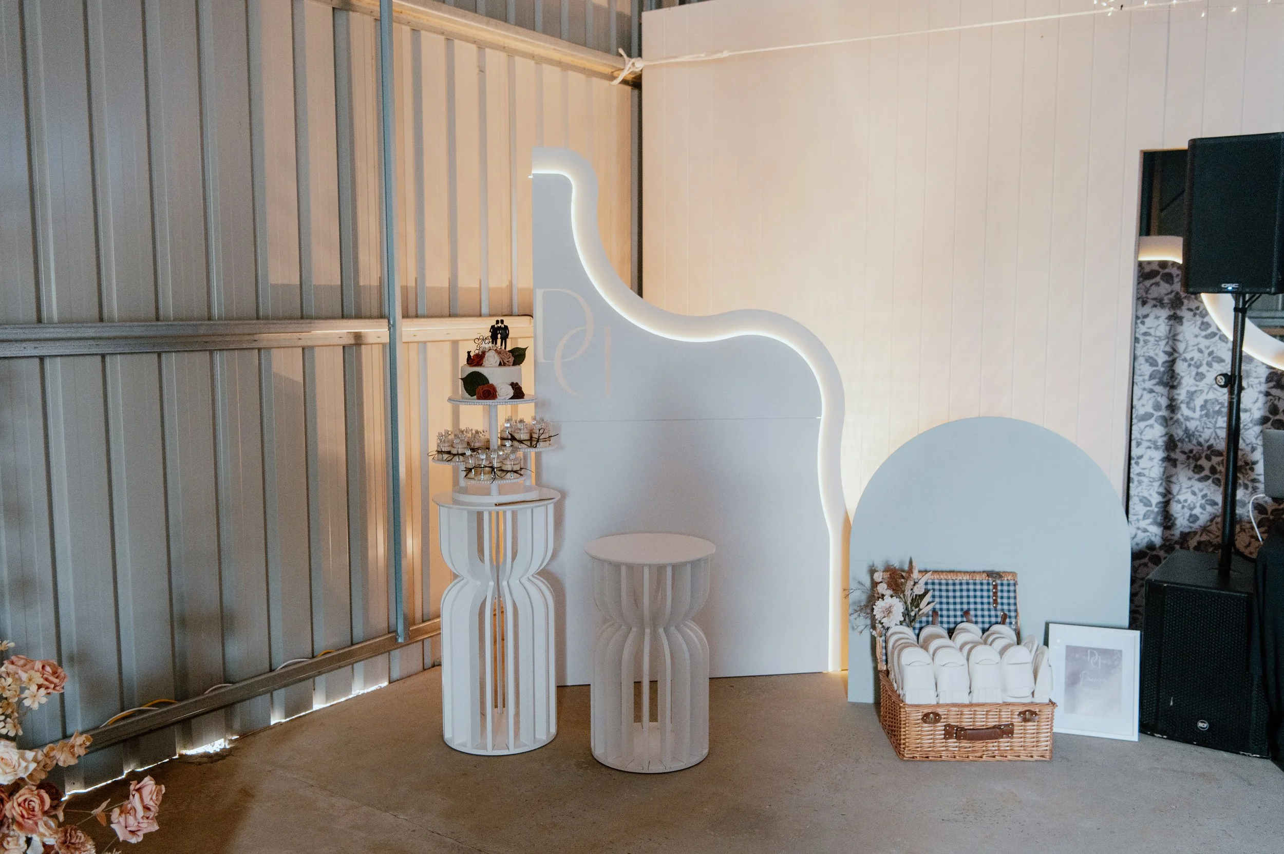 Decorative white photo booth with a curvy illuminated outline, set against a white wall with a floral curtain on the right and a metal wall on the left. The booth has a small table with supplies, and a basket holding masks or paper fans, along with f