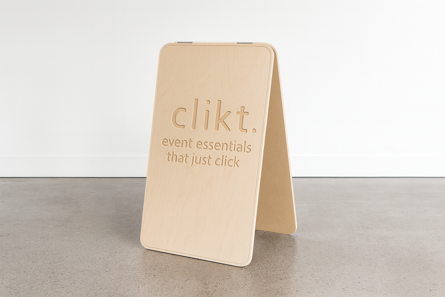 Wooden A-frame sign with engraved text that reads "clikt. event essentials that just click" placed on a concrete floor against a plain white wall.