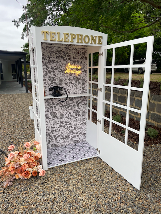 White collapsible telephone booth hire with vintage rotary phone, floral wallpaper, brass handle, mirrored ceiling, tiled base, and 'Leave a Message' LED sign—perfect for weddings, parties, and audio guestbooks in Melbourne.