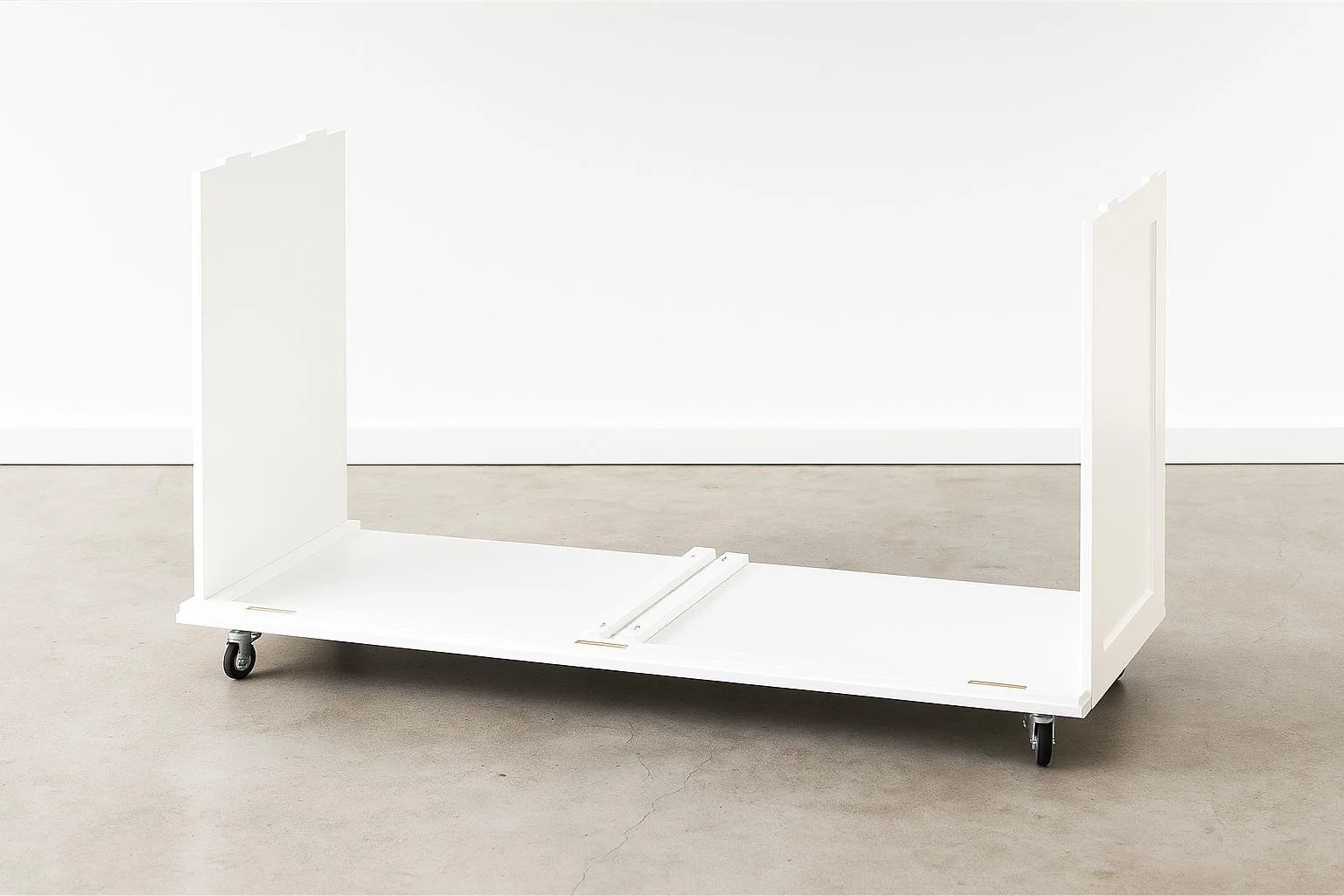 A white modular furniture piece on wheels with two large vertical panels and a flat base, possibly a room divider or display stand, set against a plain white wall.