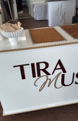A white menu or sign with the words 'TIRA' and 'MUS' displayed in large letters, placed on a white counter with a bowl of wooden stir sticks or spoons nearby.