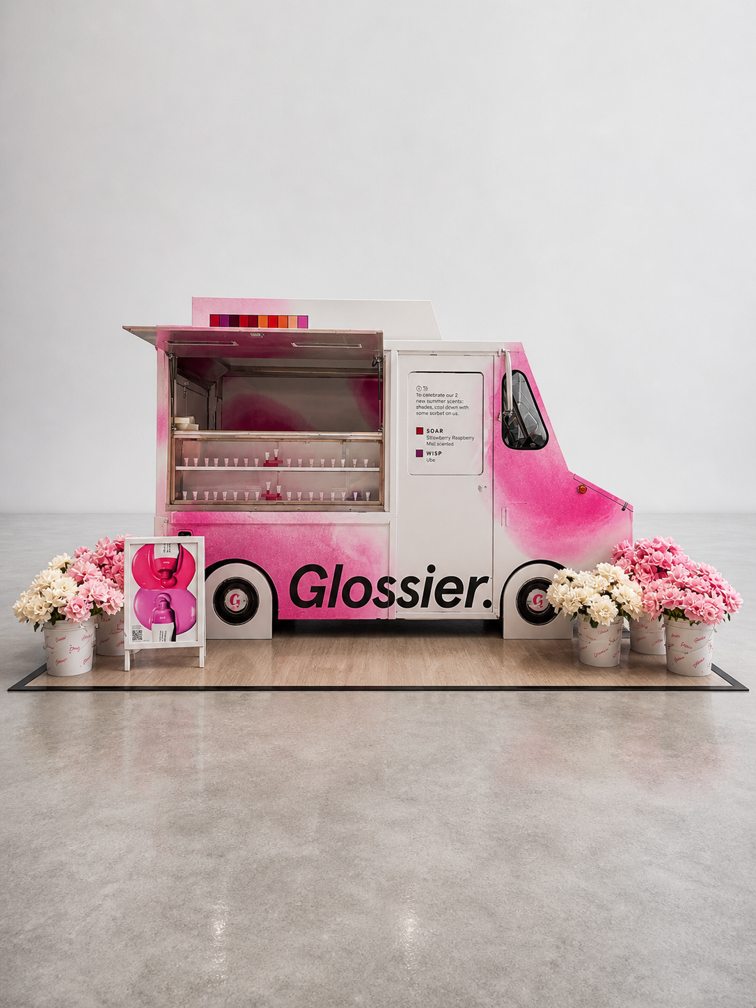 Pink mobile ice cream or snow cone van with the logo 'Glossier' on the side, surrounded by pink and white flowers, displayed in an art gallery or showroom setting.