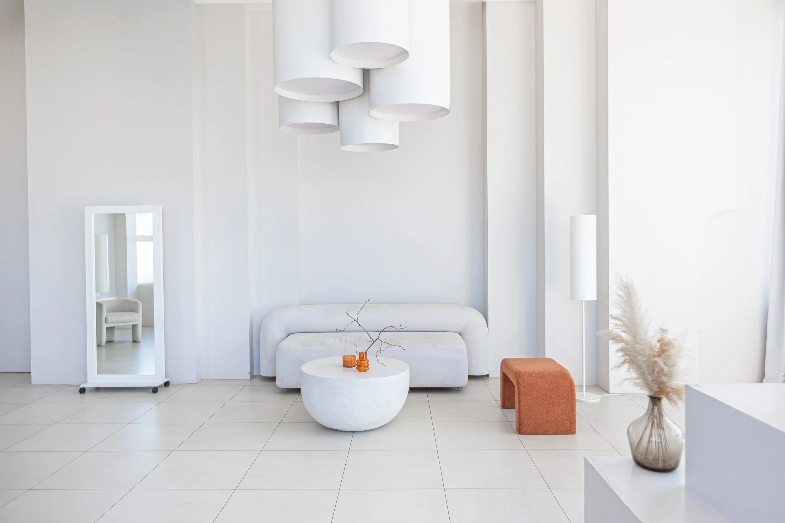 Minimalist living room with white walls and tiled floor. Contains a white sofa, round coffee table with small orange vases and bare branches, an orange ottoman, a tall white floor lamp, and a large vase with pampas grass near a white wall. A white st