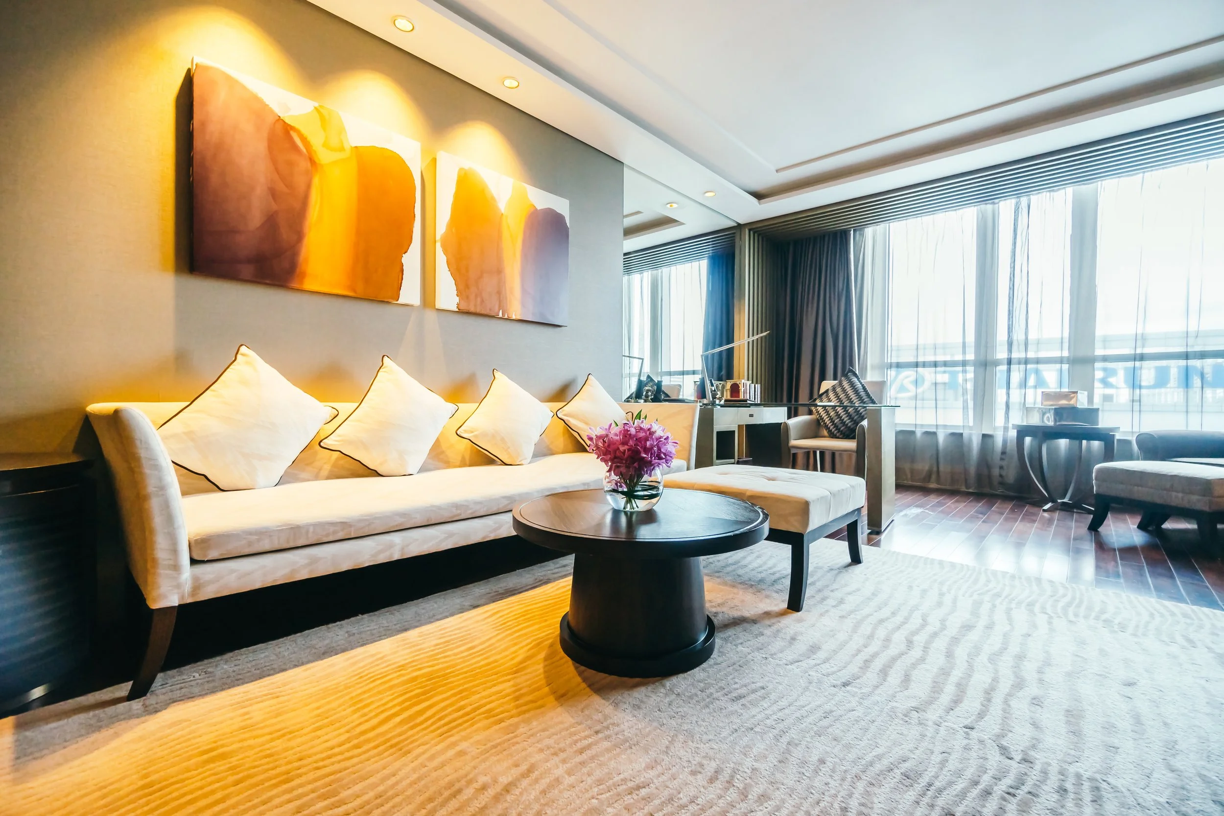 A well-lit hotel living room with a beige sofa, white pillows, a round wooden coffee table with a vase of purple flowers, a desk with a lamp, and large windows with sheer curtains and dark drapes.