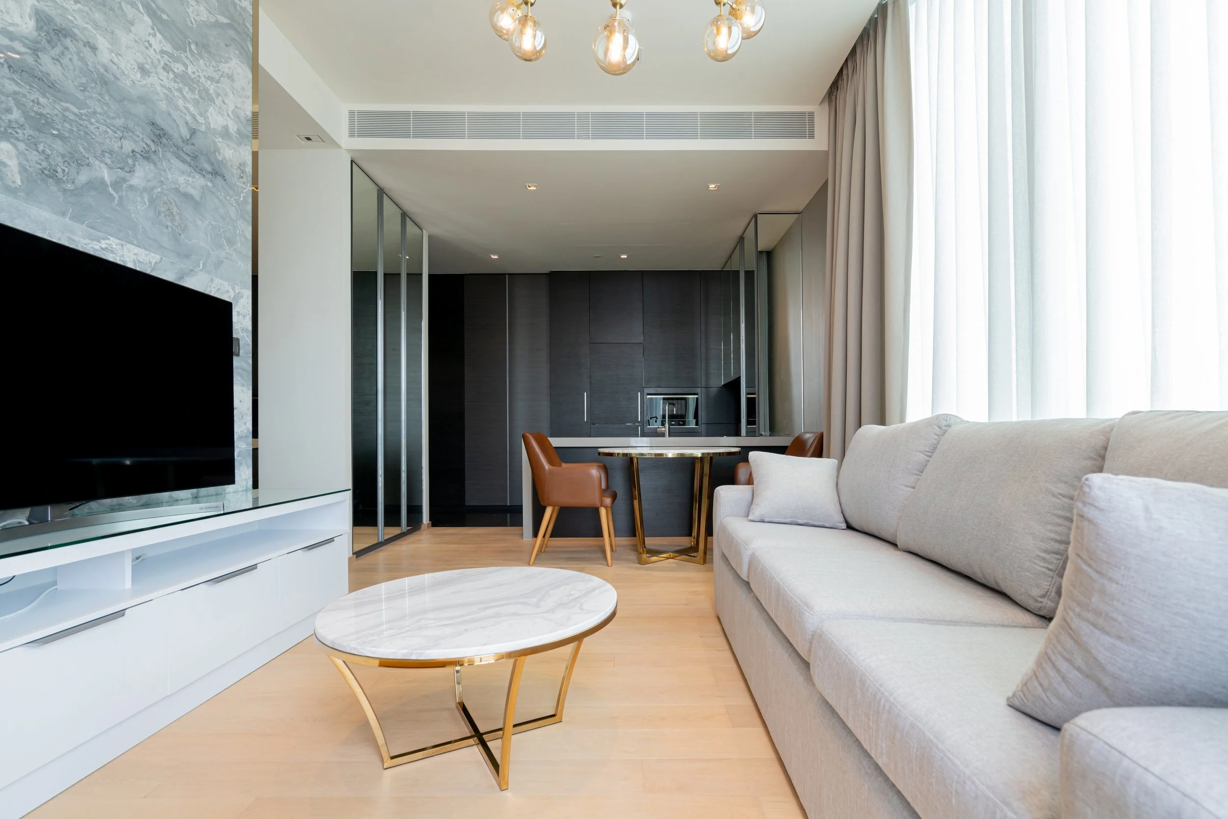 Modern living room with beige sofa, marble-top coffee table, TV on white media console, and large window with white curtains. In the background, a dining area with leather chairs and a dark wall kitchen is visible, with ceiling lights.