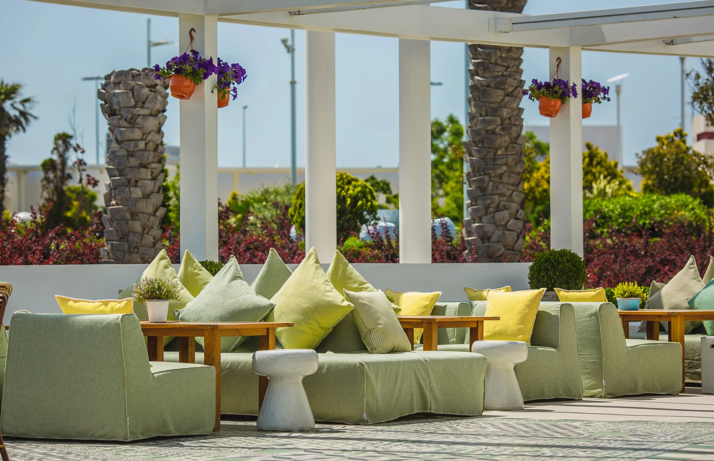 Outdoor seating area with lime green cushioned sofas and pillows, small potted plants on tables, white pillars, purple hanging flowers, palm trees, and lush greenery in the background.