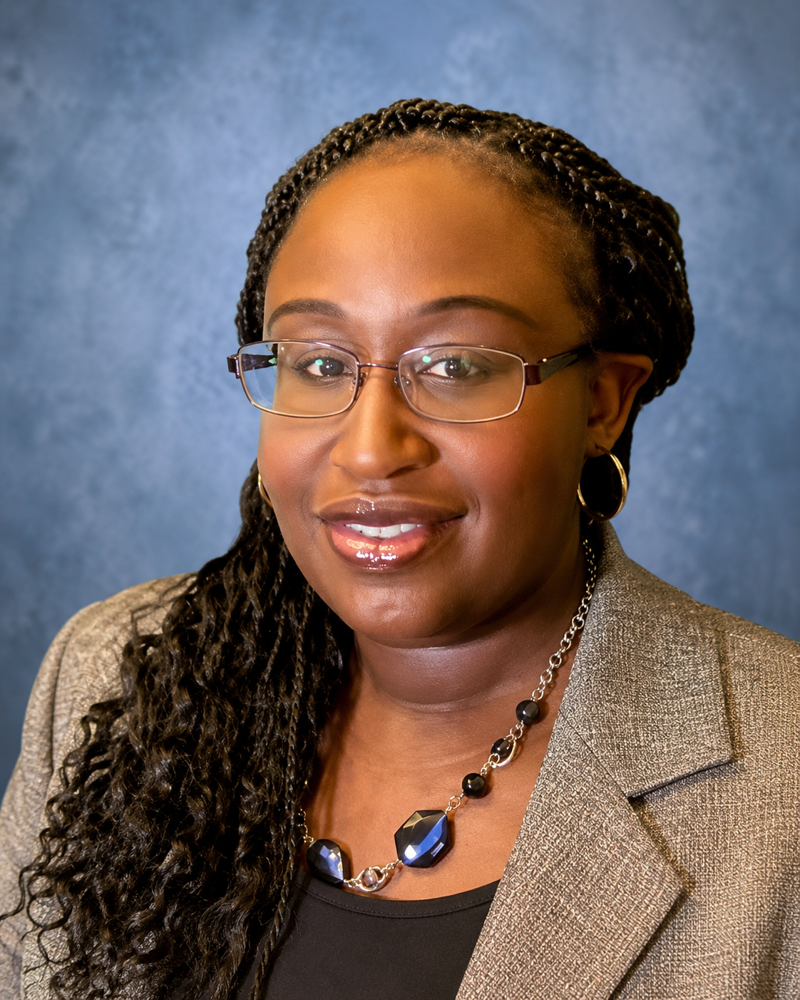 Portrait of a woman wearing glasses, earrings, and a beige blazer, with a neutral background.